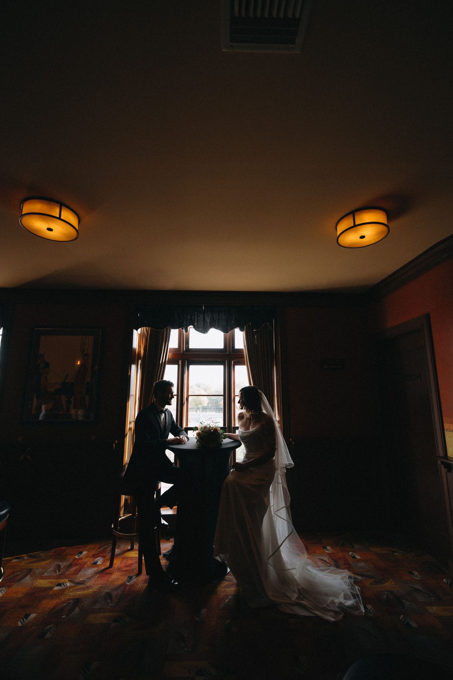 Silhouette of bride and groom sitting at table by window in warmly lit vintage room.