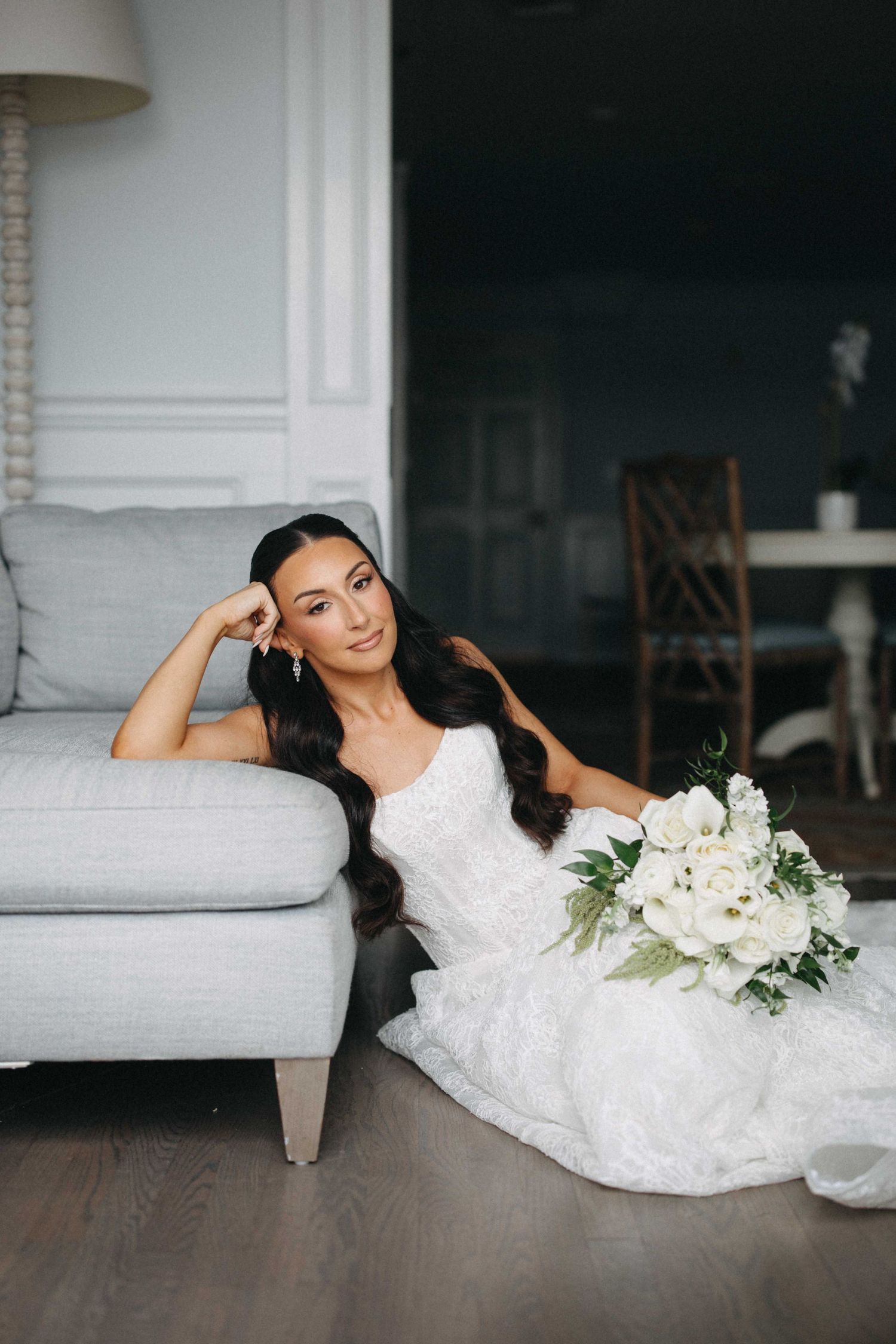 Bride in white lace gown sits on floor leaning against gray sofa holding white floral bouquet.