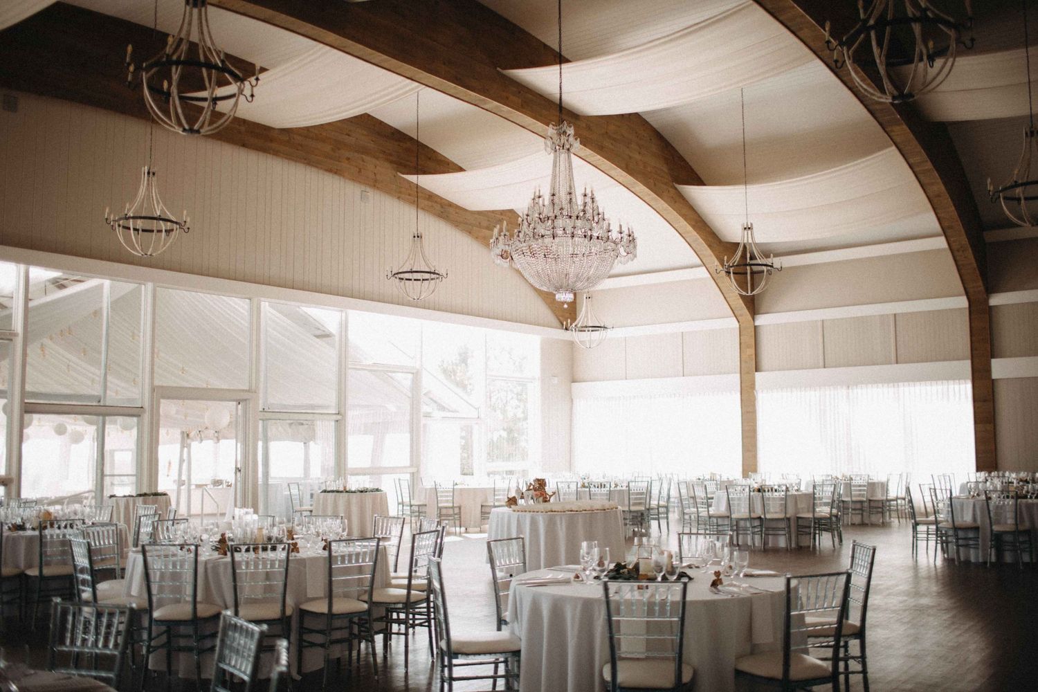 Elegant wedding reception hall with crystal chandeliers, wooden beams, and round draped tables.