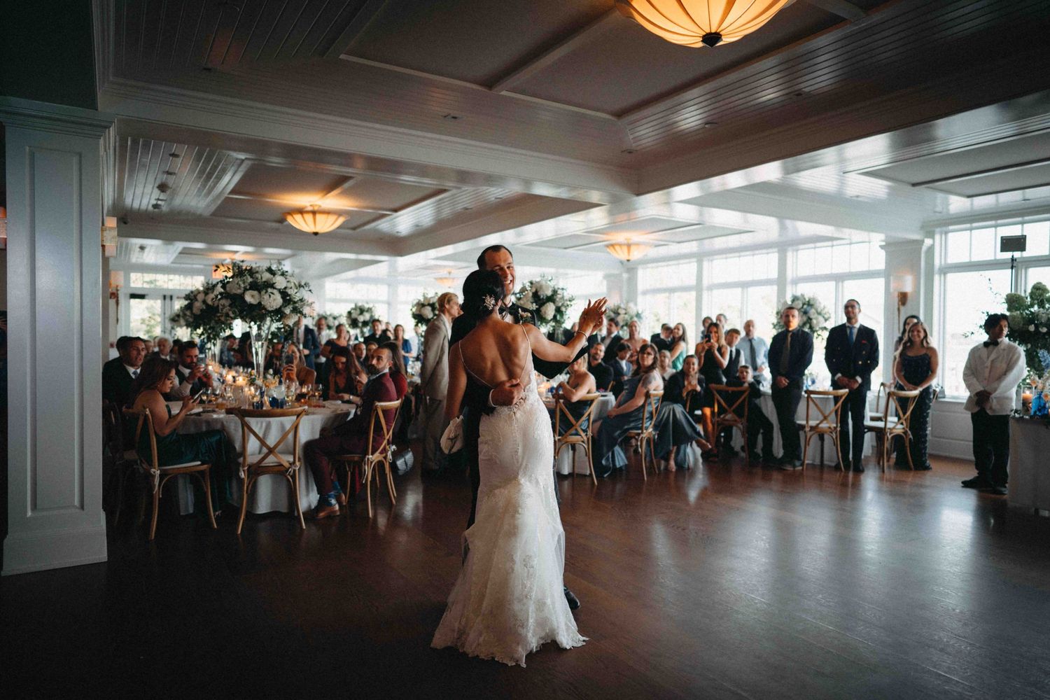 Bride in backless lace wedding gown addressing seated wedding reception guests in waterfront venue.