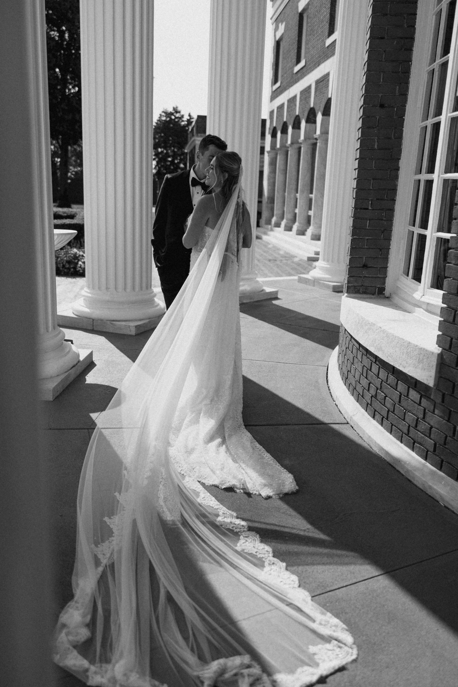 Black and white photo of bride and groom kissing among white columns, brides long lace veil trailing elegantly.