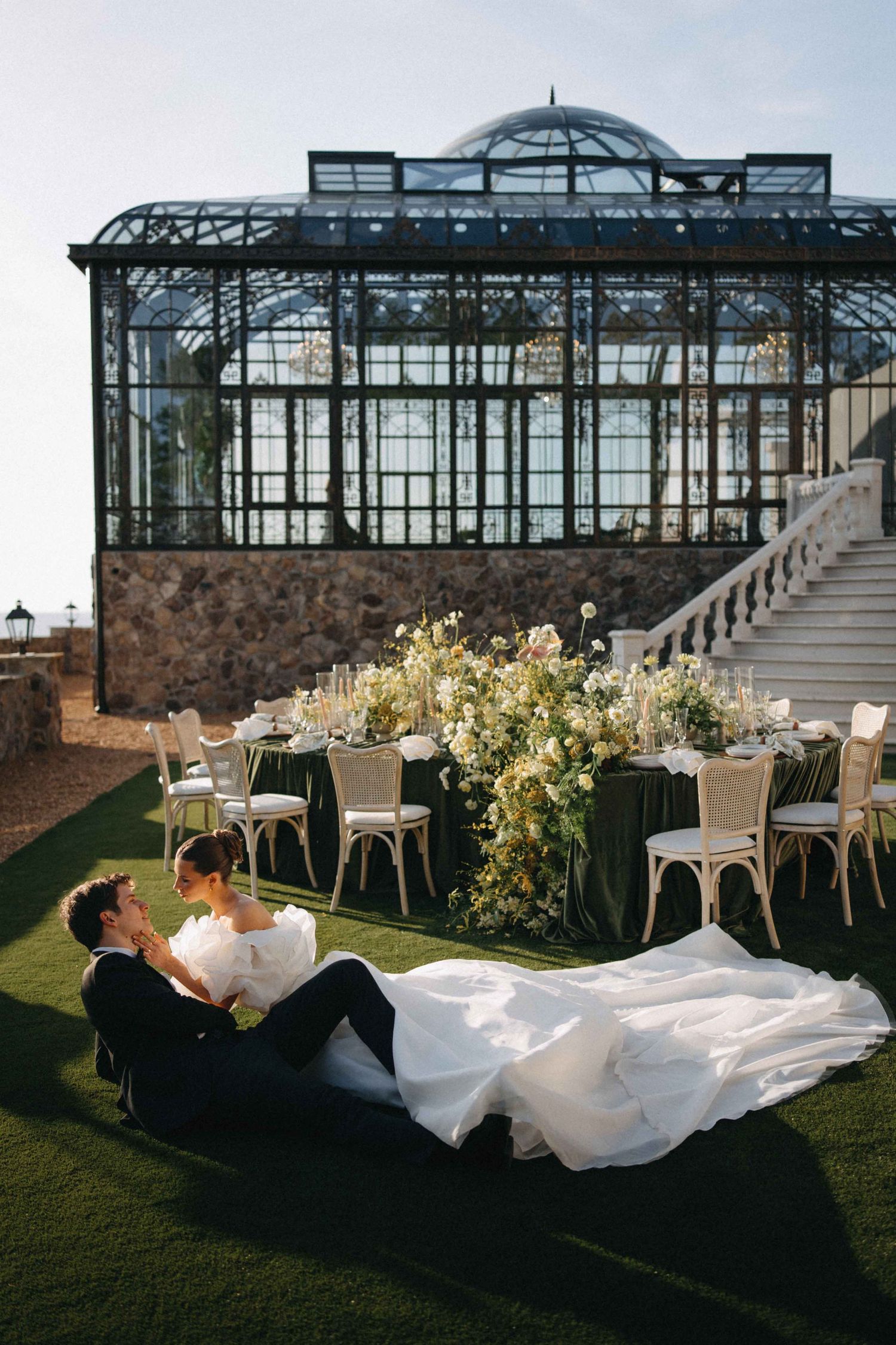 Bride and groom relaxing on lawn before elegant glass greenhouse wedding reception with floral centerpieces.