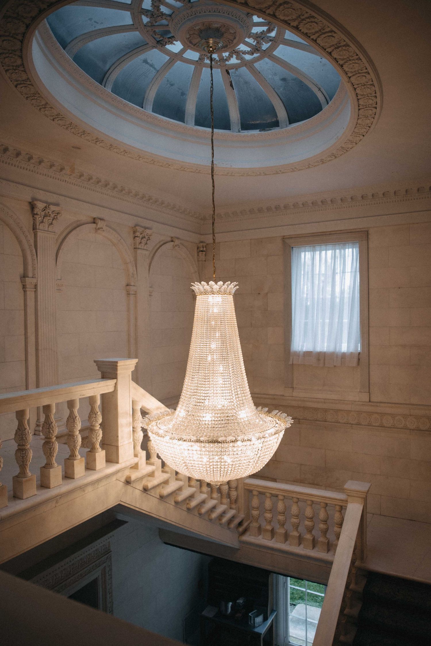 Stunning empire crystal chandelier hangs beneath circular skylight dome in ornate neoclassical venue with balustrade railing.