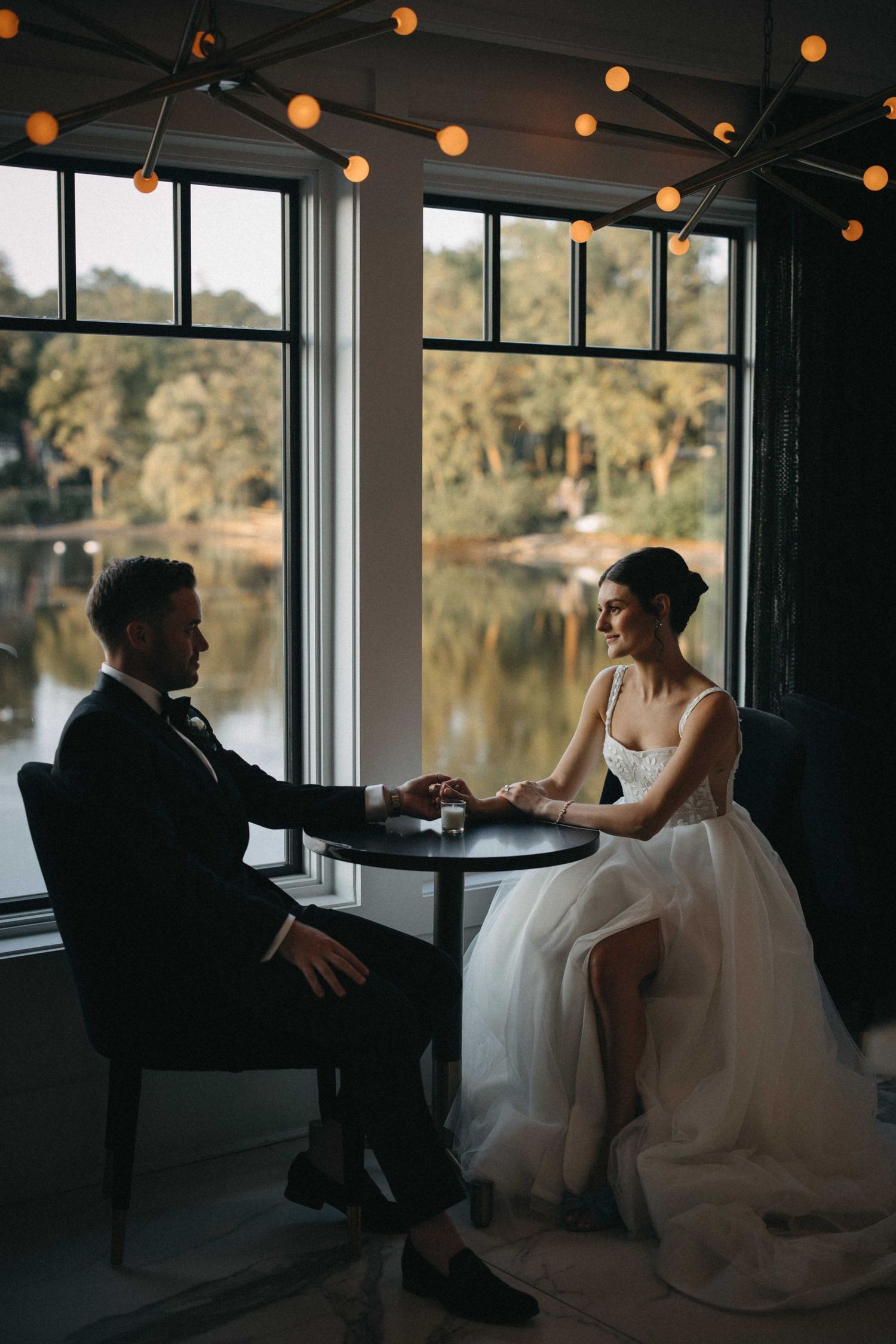 Bride and groom holding hands at intimate table by waterfront window with string lights.