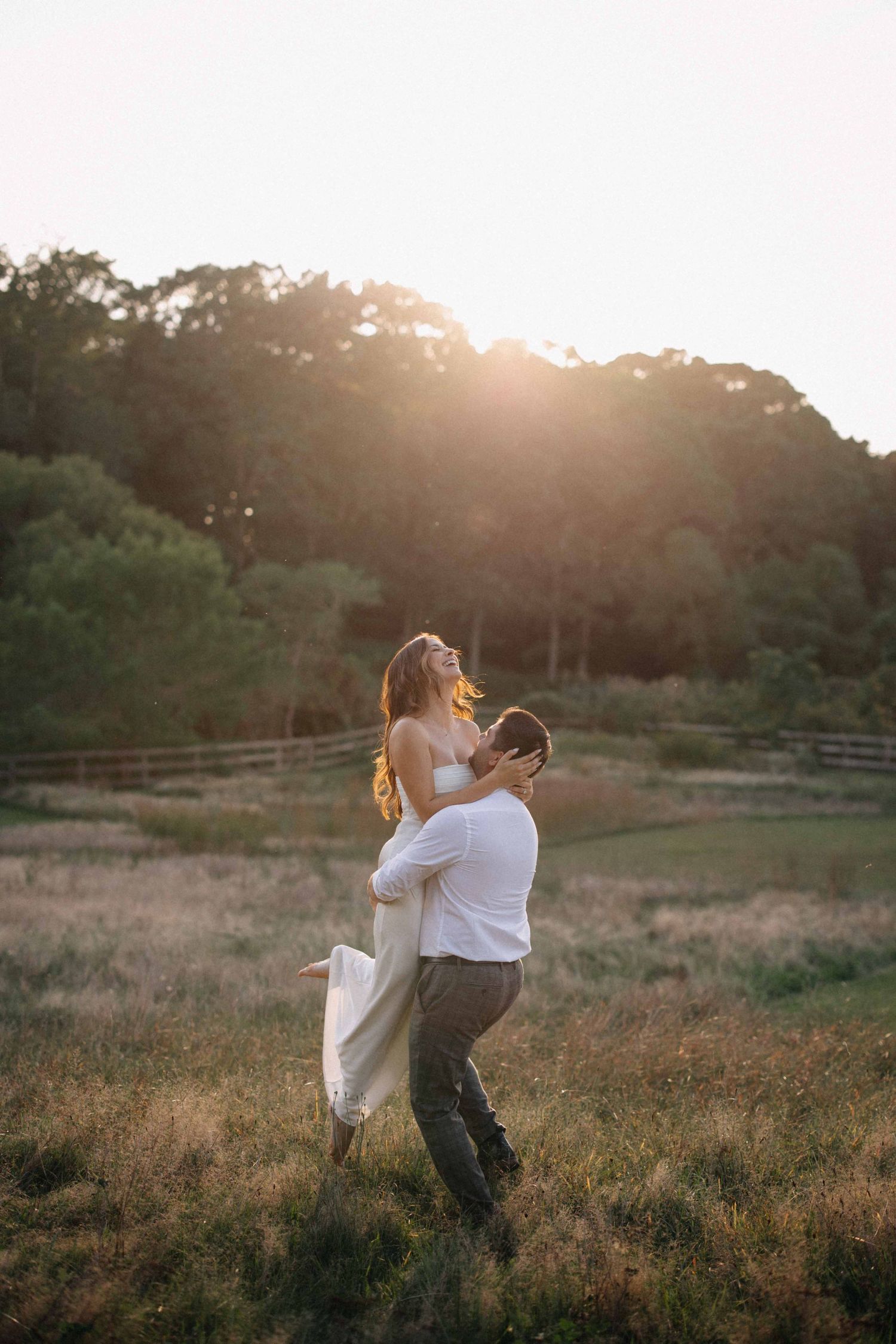 Man lifting woman in white dress at golden sunset in open field with wooden fence and trees.