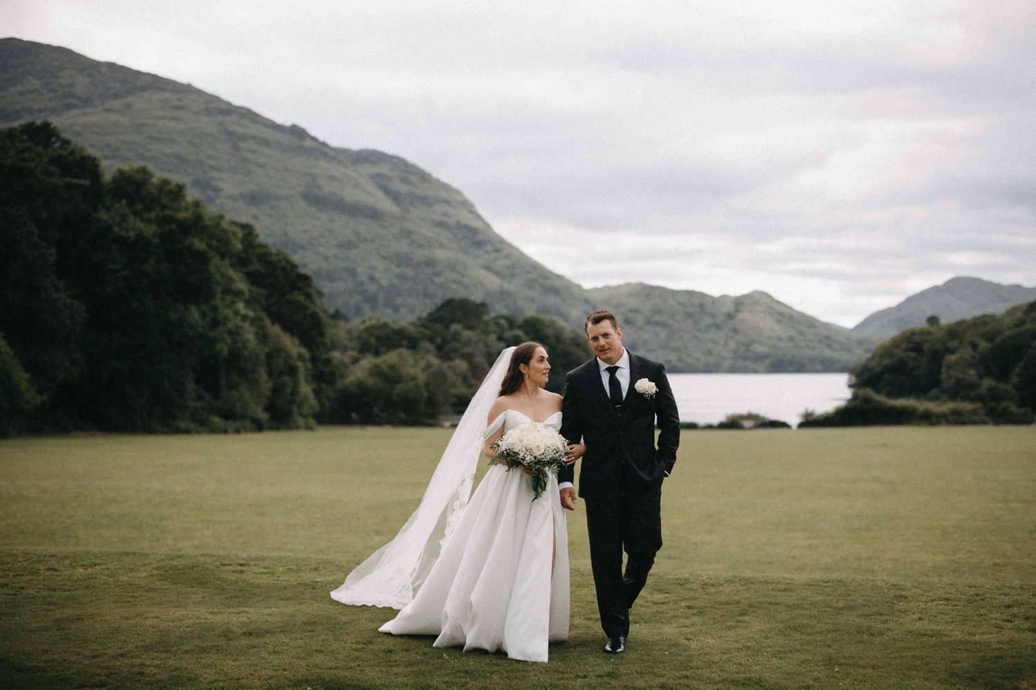 Bride and groom walk hand in hand across green meadow with scenic mountain lake backdrop on wedding day.