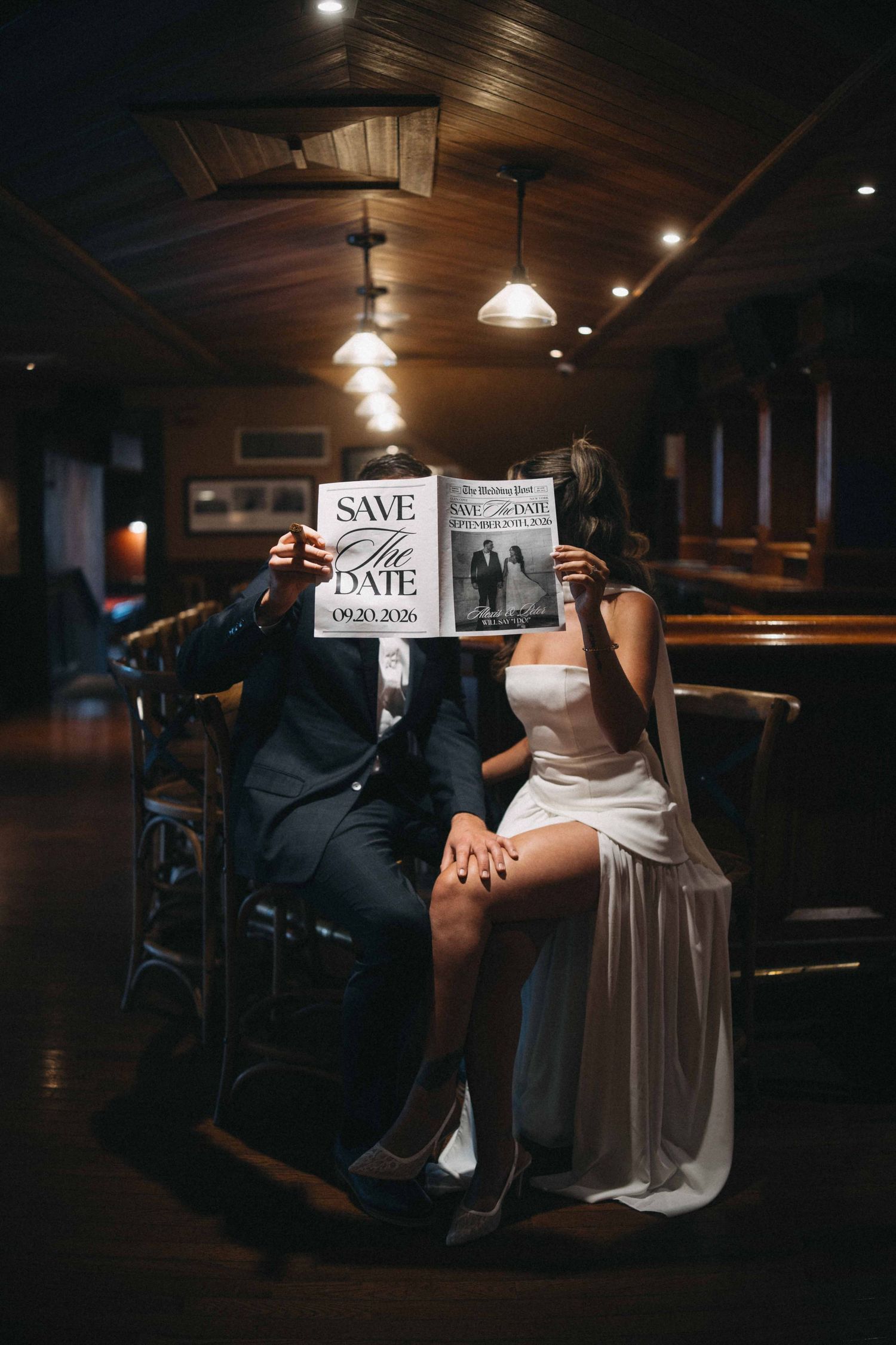 Engaged couple hides faces behind Save the Date cards in dimly lit bar.
