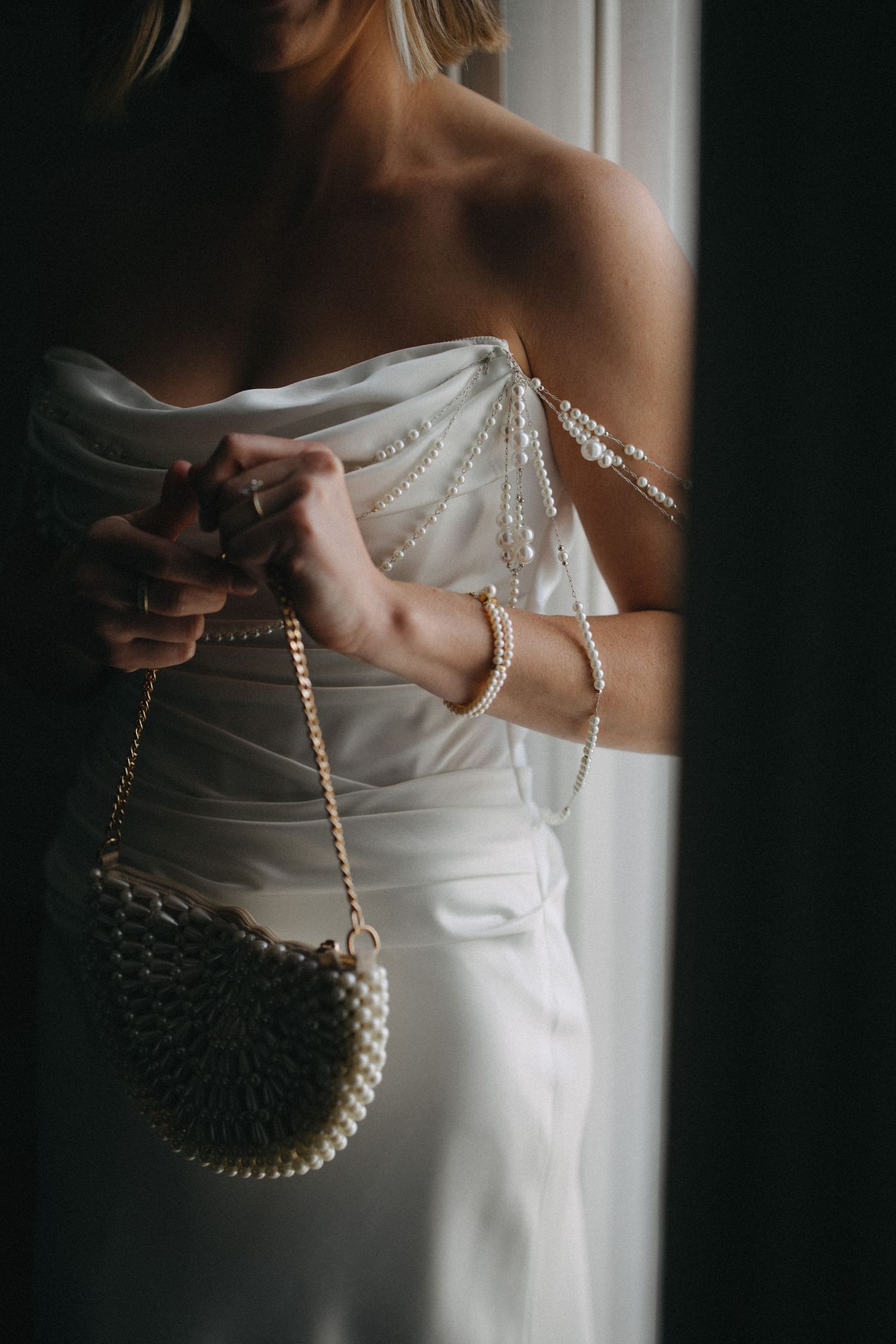 Bride in strapless white gown holding pearl clutch bag with pearl strap details by window.