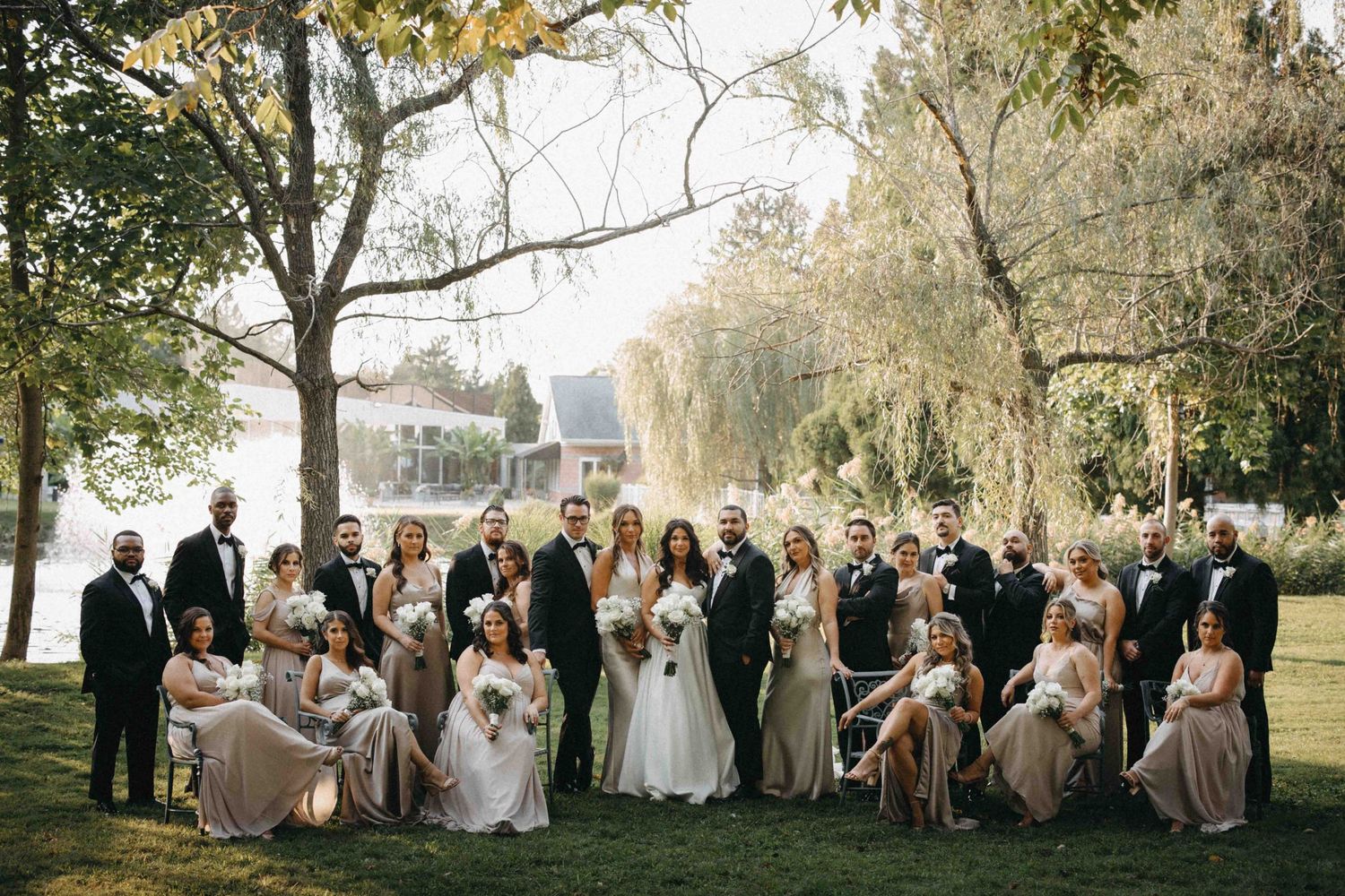 Wedding party in neutral gowns and black tuxedos poses on lush green lawn near willow tree.