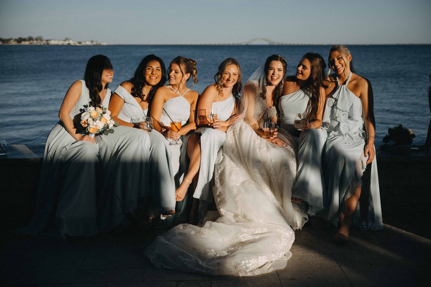 Bride and six bridesmaids in blue gowns laughing together while seated waterside in golden hour light.