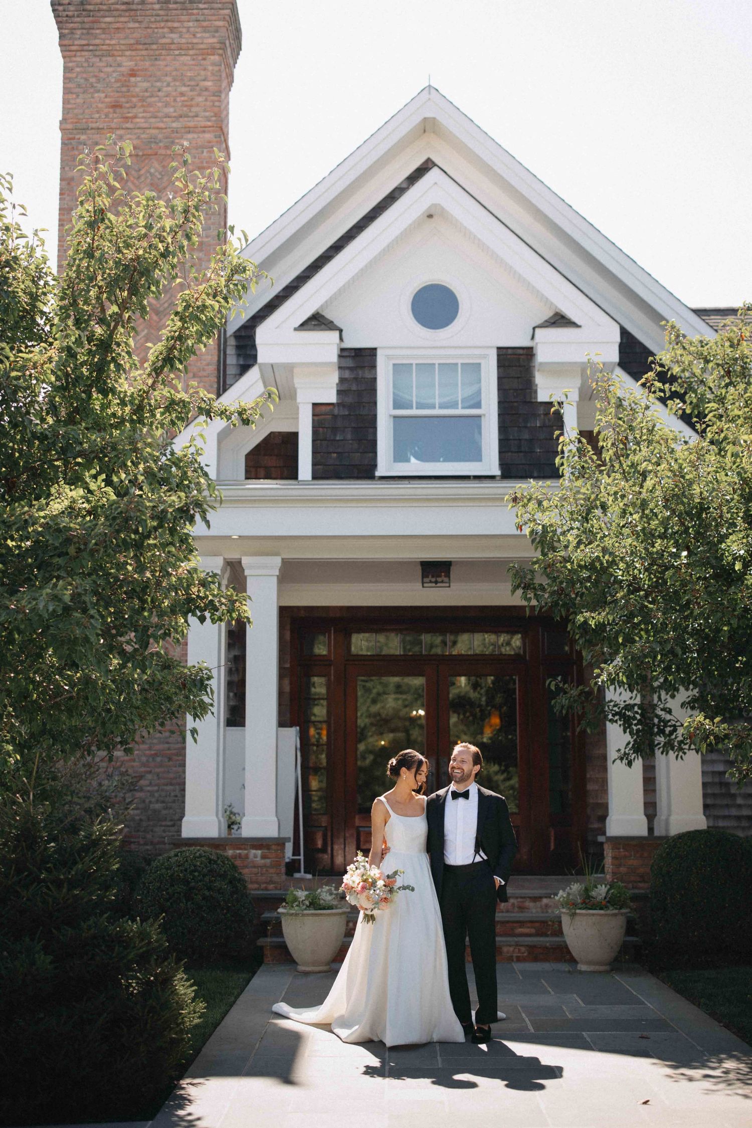 Bride in white gown and groom in black tuxedo stand before a charming brick and shingle wedding venue.