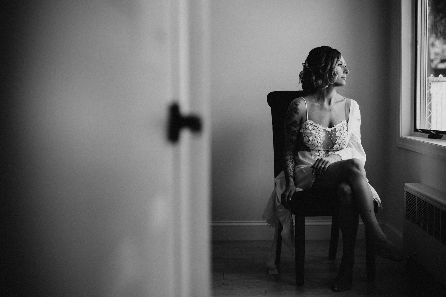 Black and white photo of bride in lace robe sitting thoughtfully by a sunlit window.