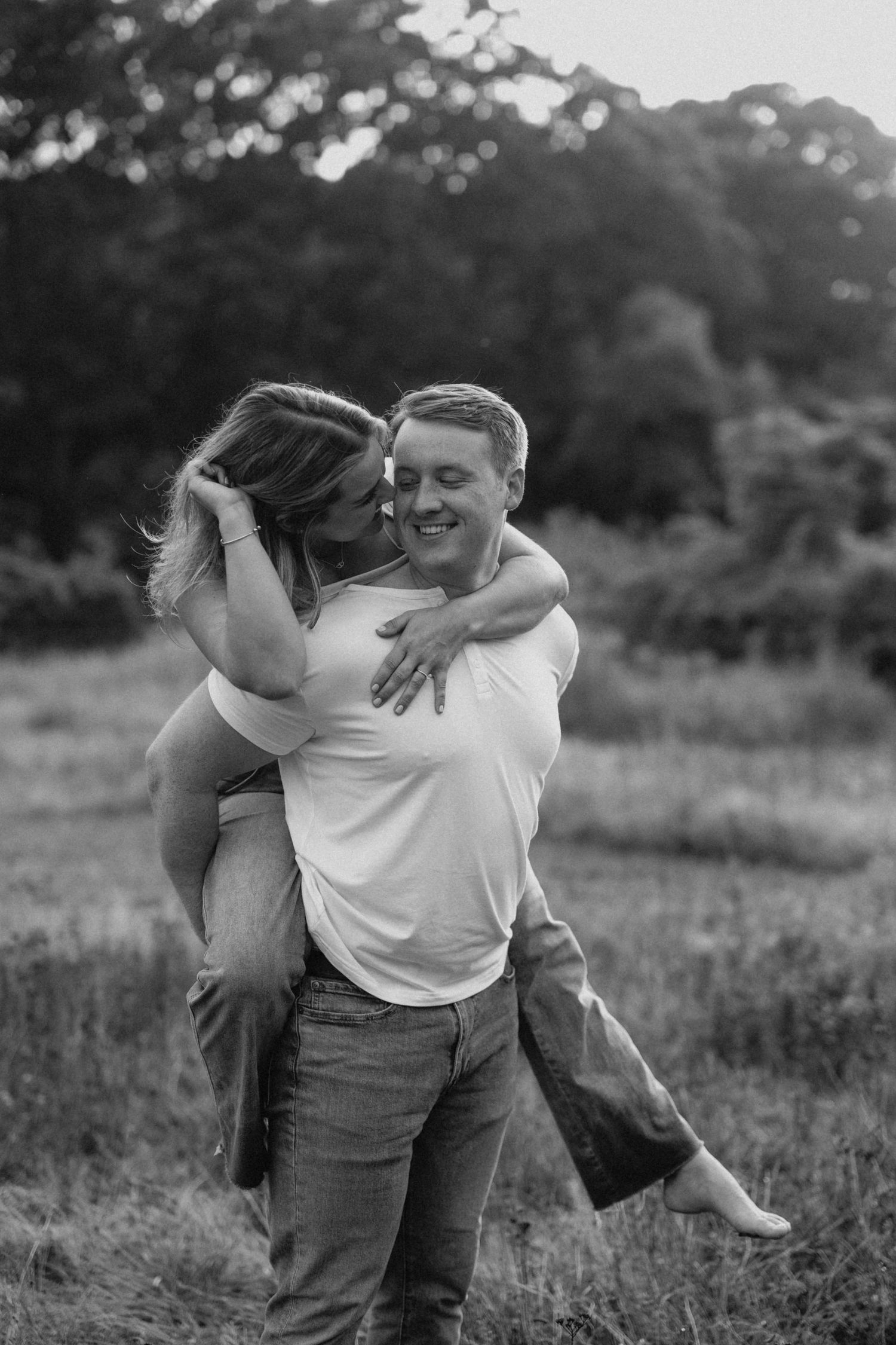 Black and white photo of a smiling man giving his laughing partner a piggyback ride in a meadow.
