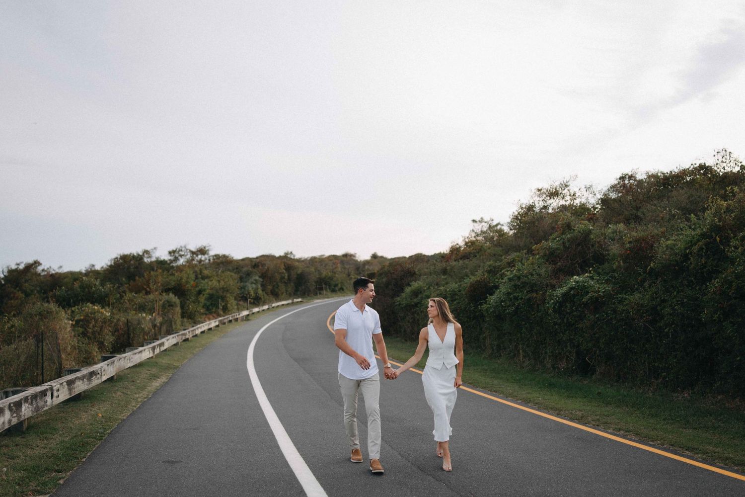 Couple holding hands walking down a winding road surrounded by lush greenery.