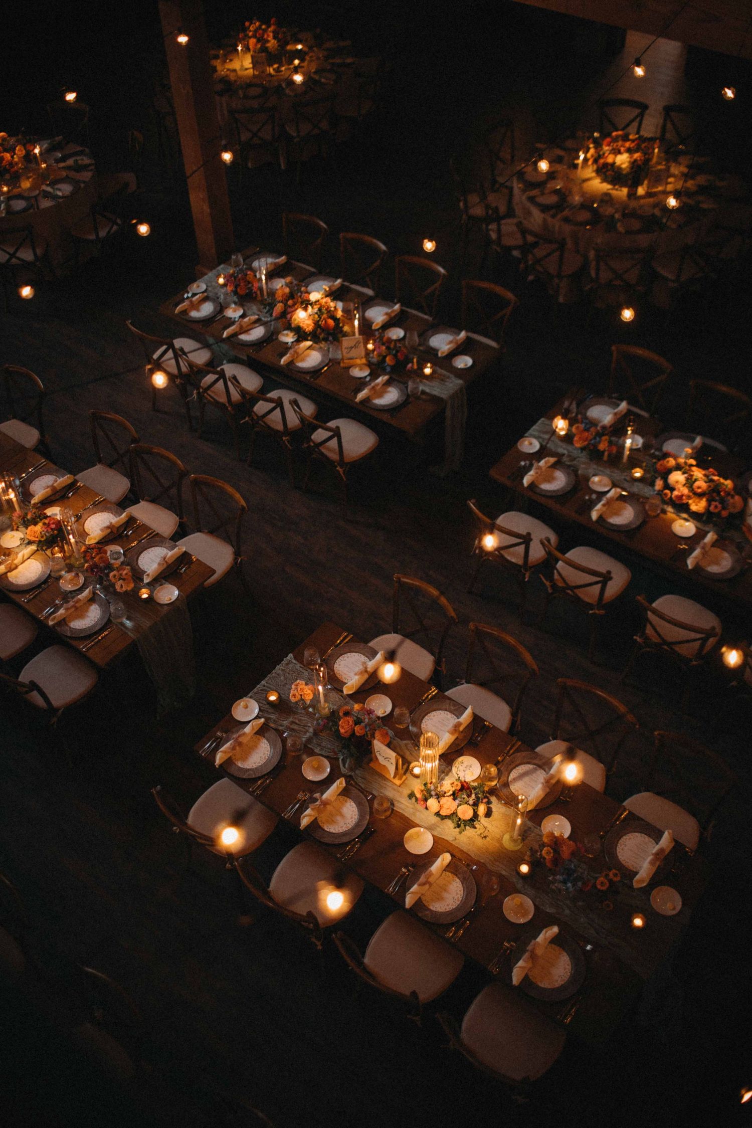 Aerial view of elegantly set wedding reception tables adorned with candles and floral centerpieces in a dark barn.