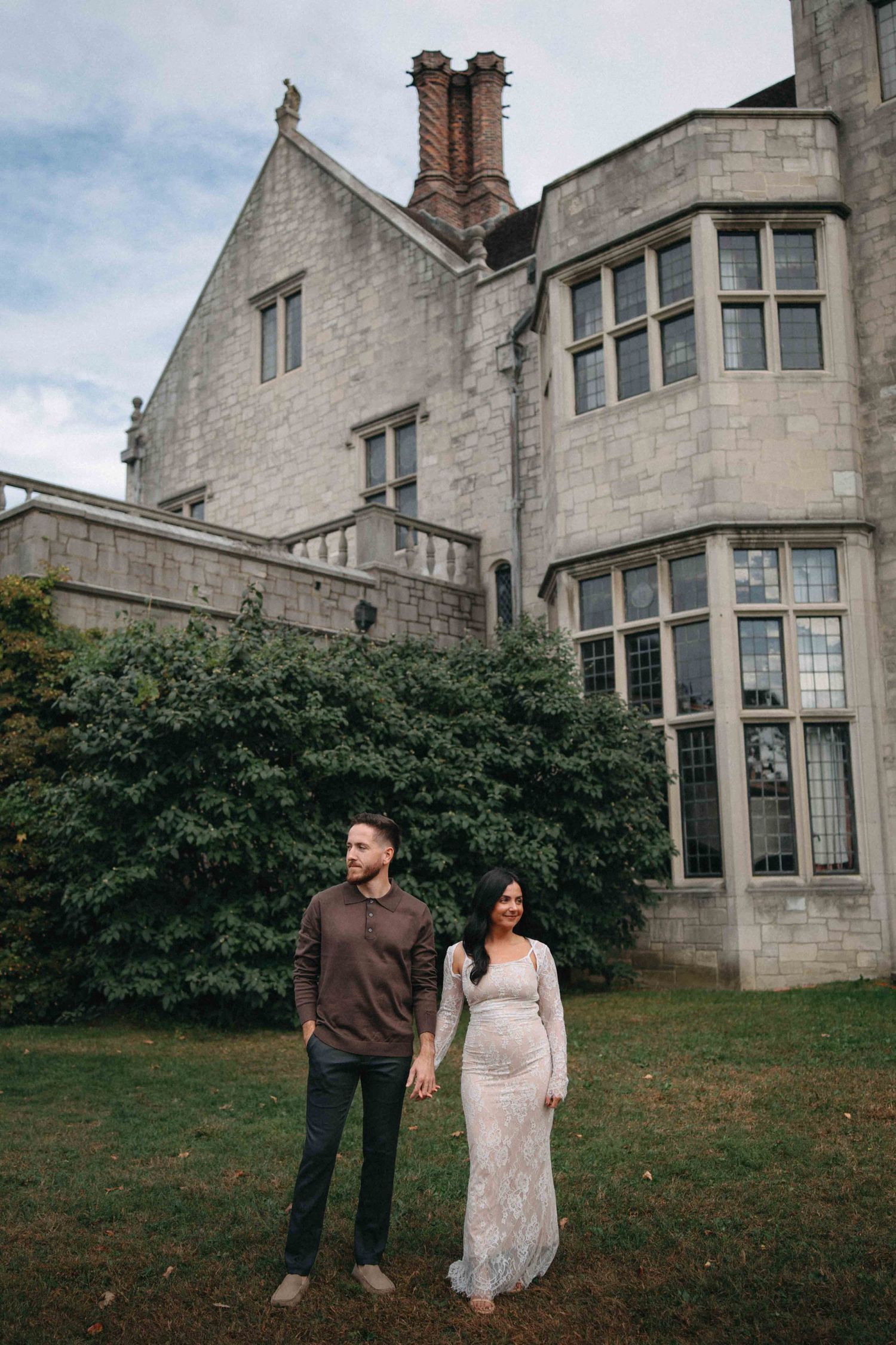 Couple holding hands on lawn in front of a grand stone manor house.