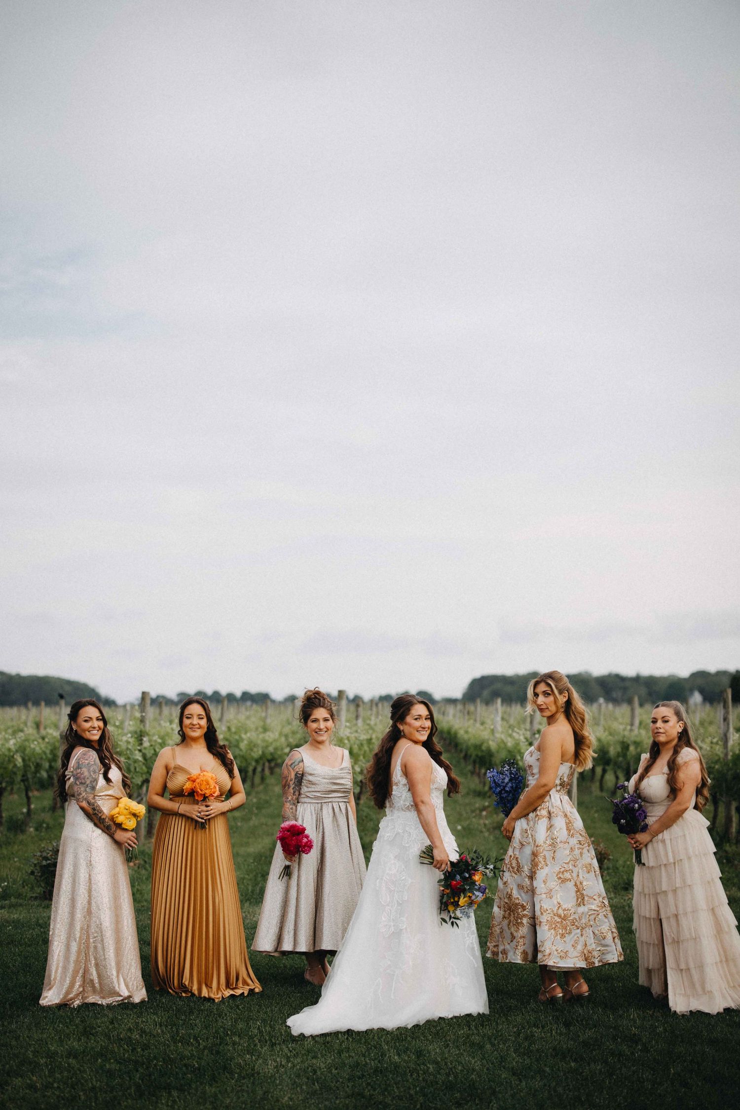 Bride with bridesmaids in mismatched colorful dresses pose in front of vineyard rows.