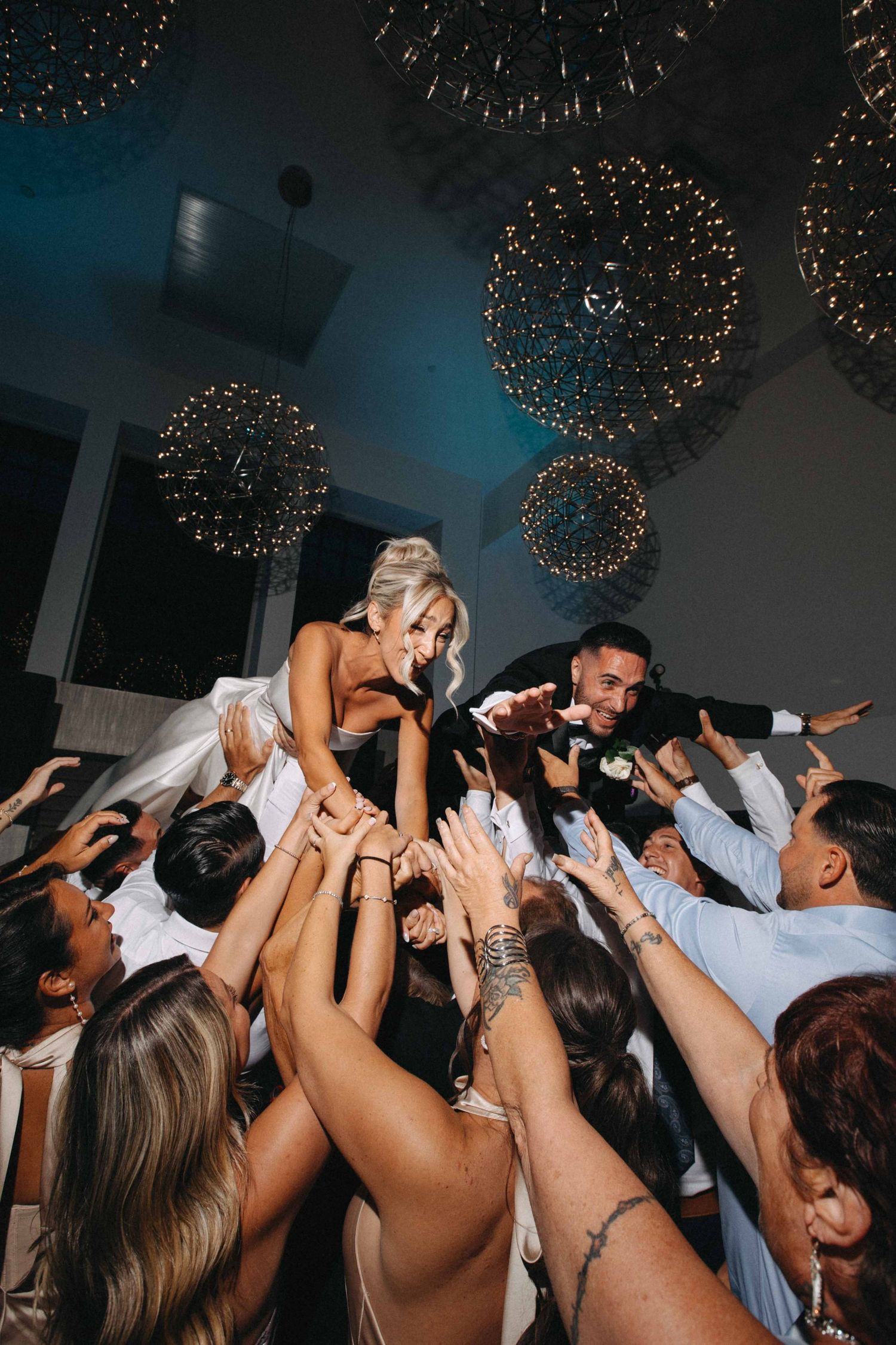 Bride being lifted by wedding guests during lively reception party under sparkling chandelier lights.