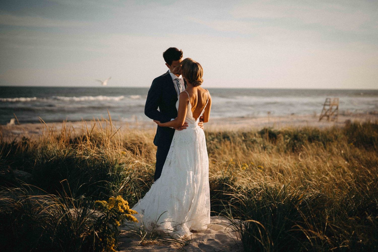 Bride and groom embracing in tall beach grass with ocean waves in the background.
