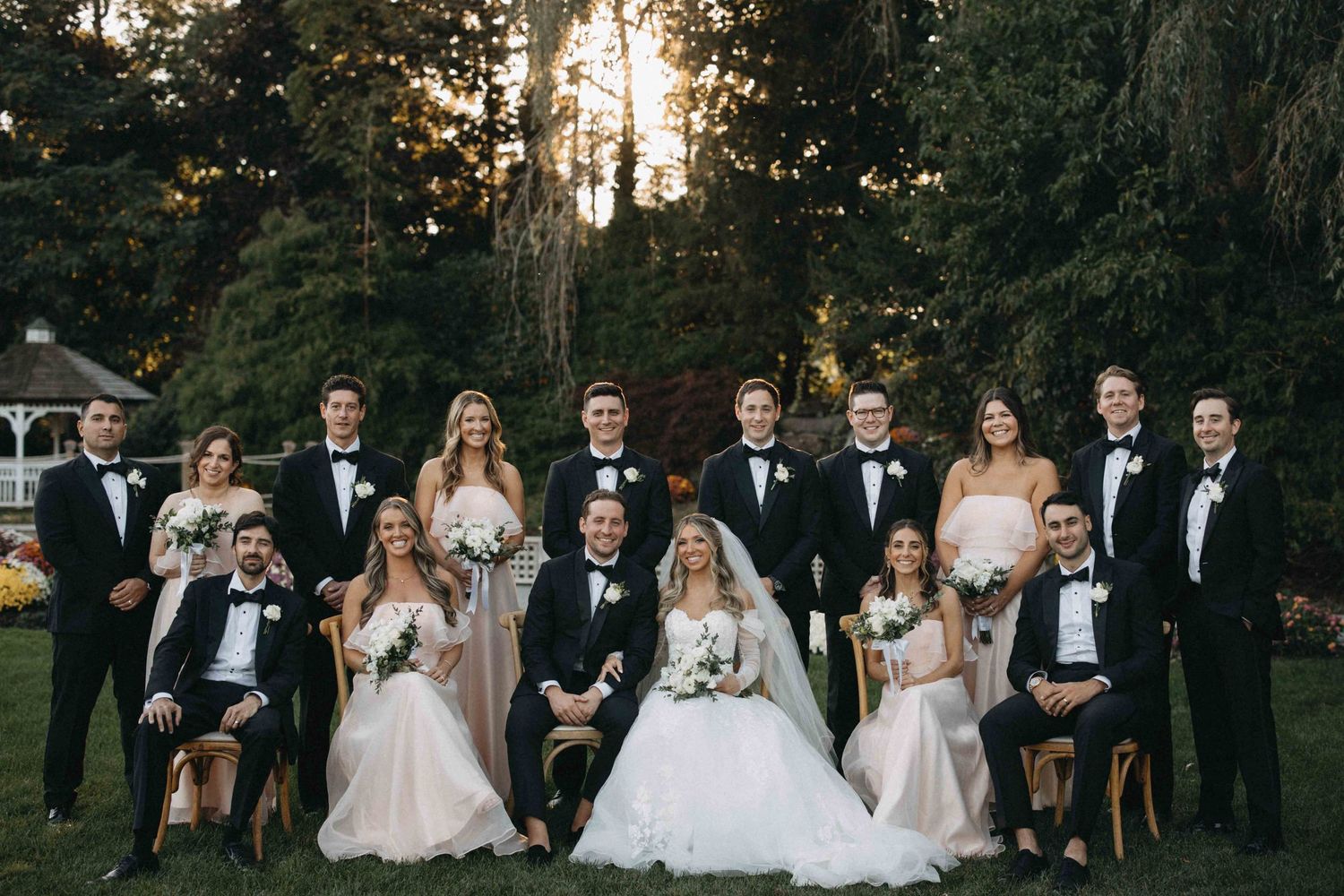 Large wedding party posing together in formal attire at sunset garden venue.