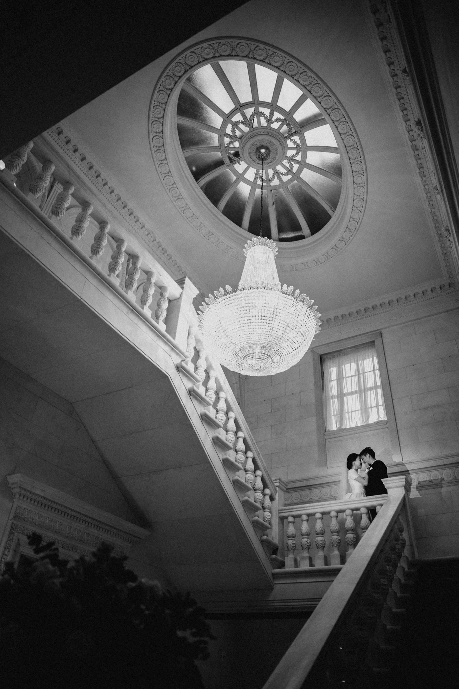 Black and white photo of a couple kissing on a grand staircase beneath a crystal chandelier.