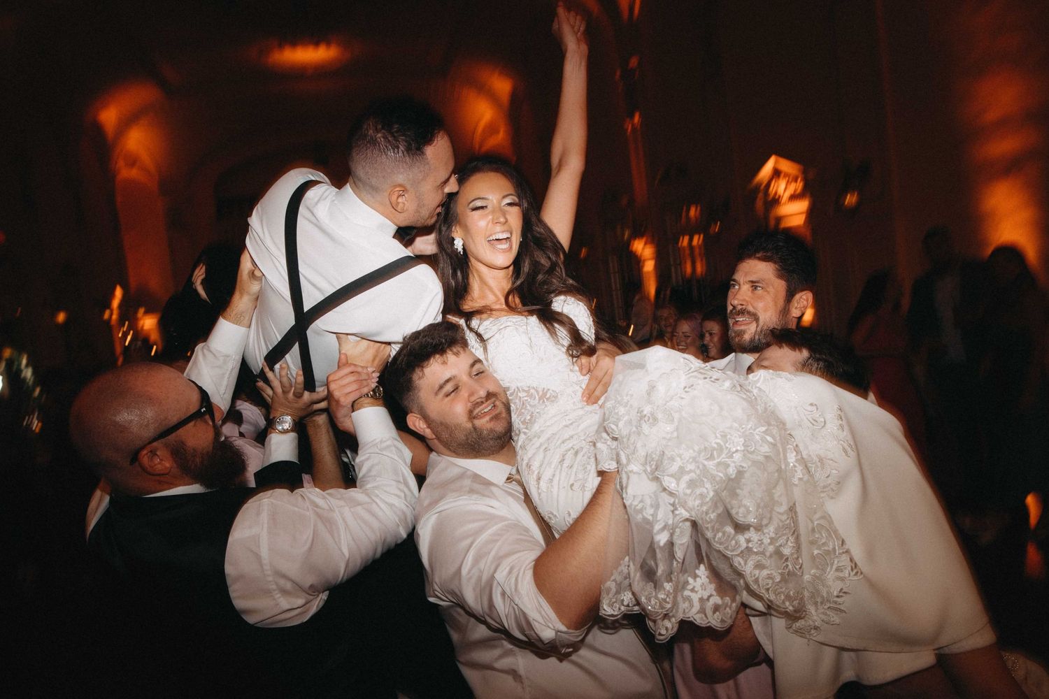 Joyful bride lifted by groomsmen during energetic wedding reception celebration with crowd cheering around them.