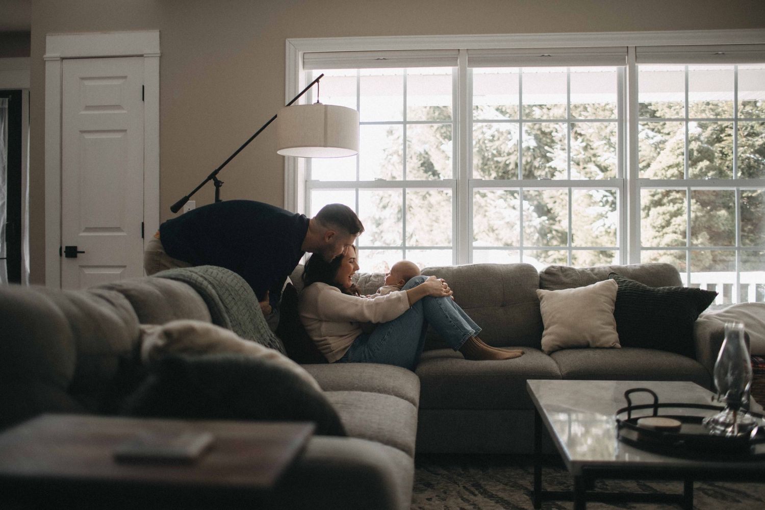 Couple sharing a romantic moment on a gray sectional sofa in a cozy living room with large windows.