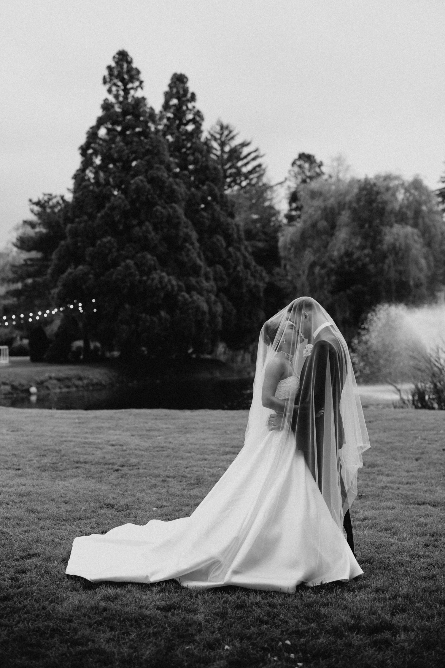 Black and white photo of a bride in a flowing gown and veil standing on a lawn with trees in the background.