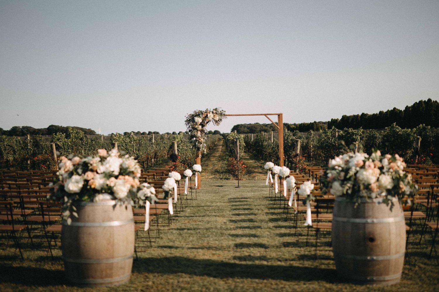 Outdoor vineyard wedding ceremony aisle with barrel florals and wooden arch at sunset.