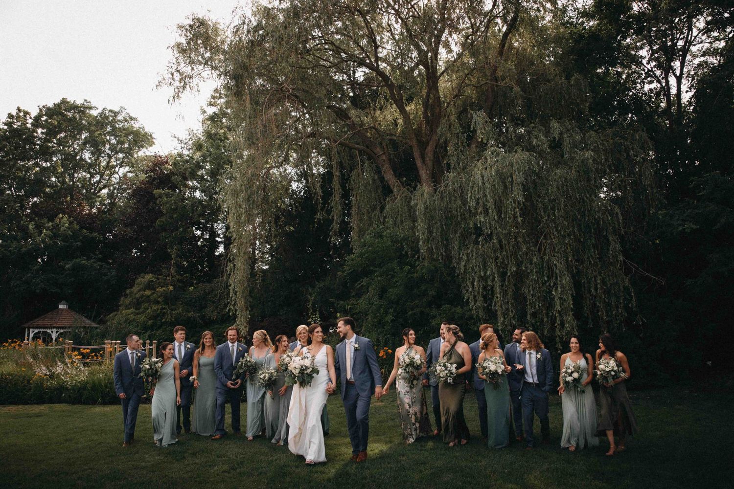 Wedding party group photo on lush green lawn beneath a weeping willow tree at twilight.