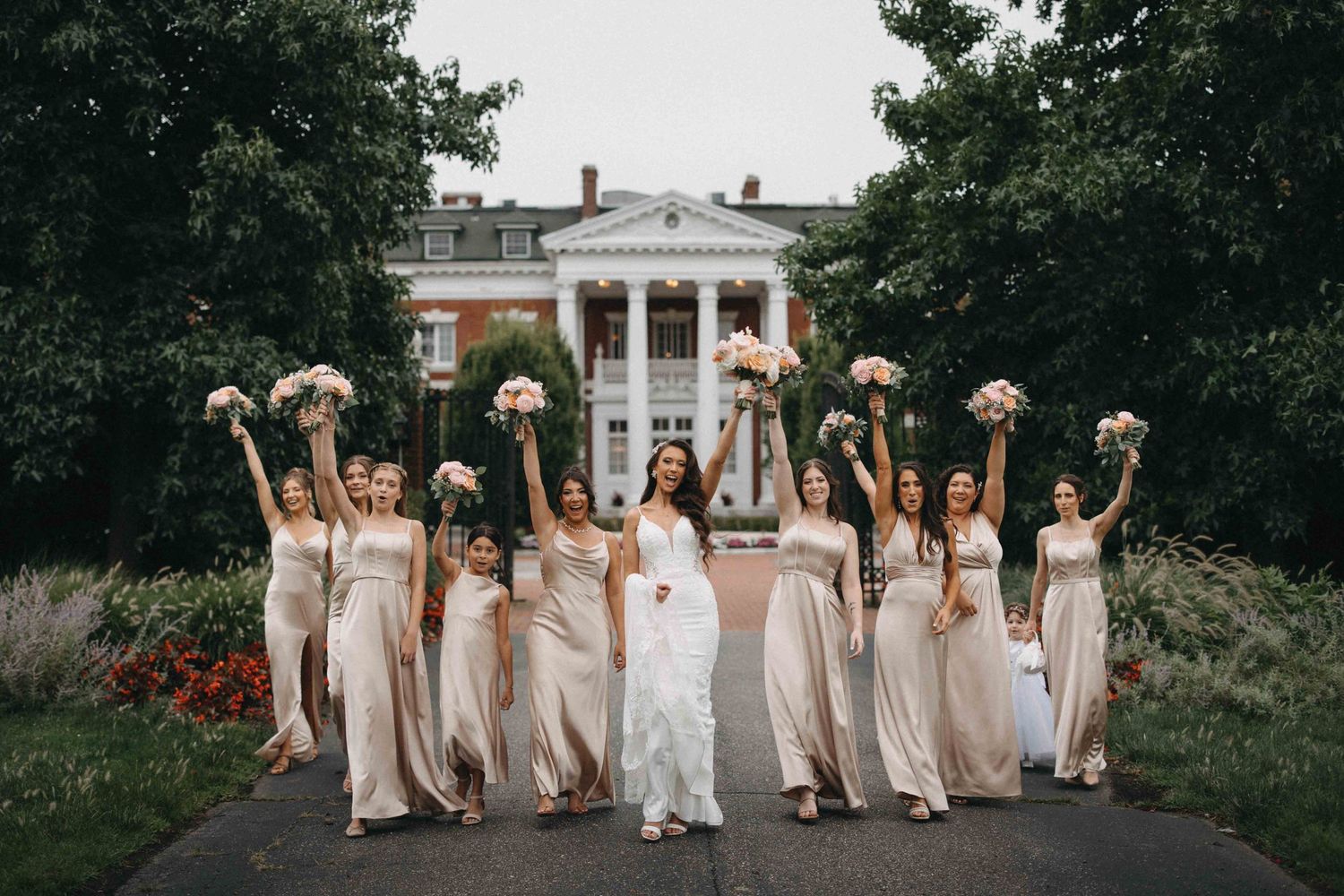 Bride and bridesmaids in champagne dresses joyfully raise bouquets before white colonial mansion.