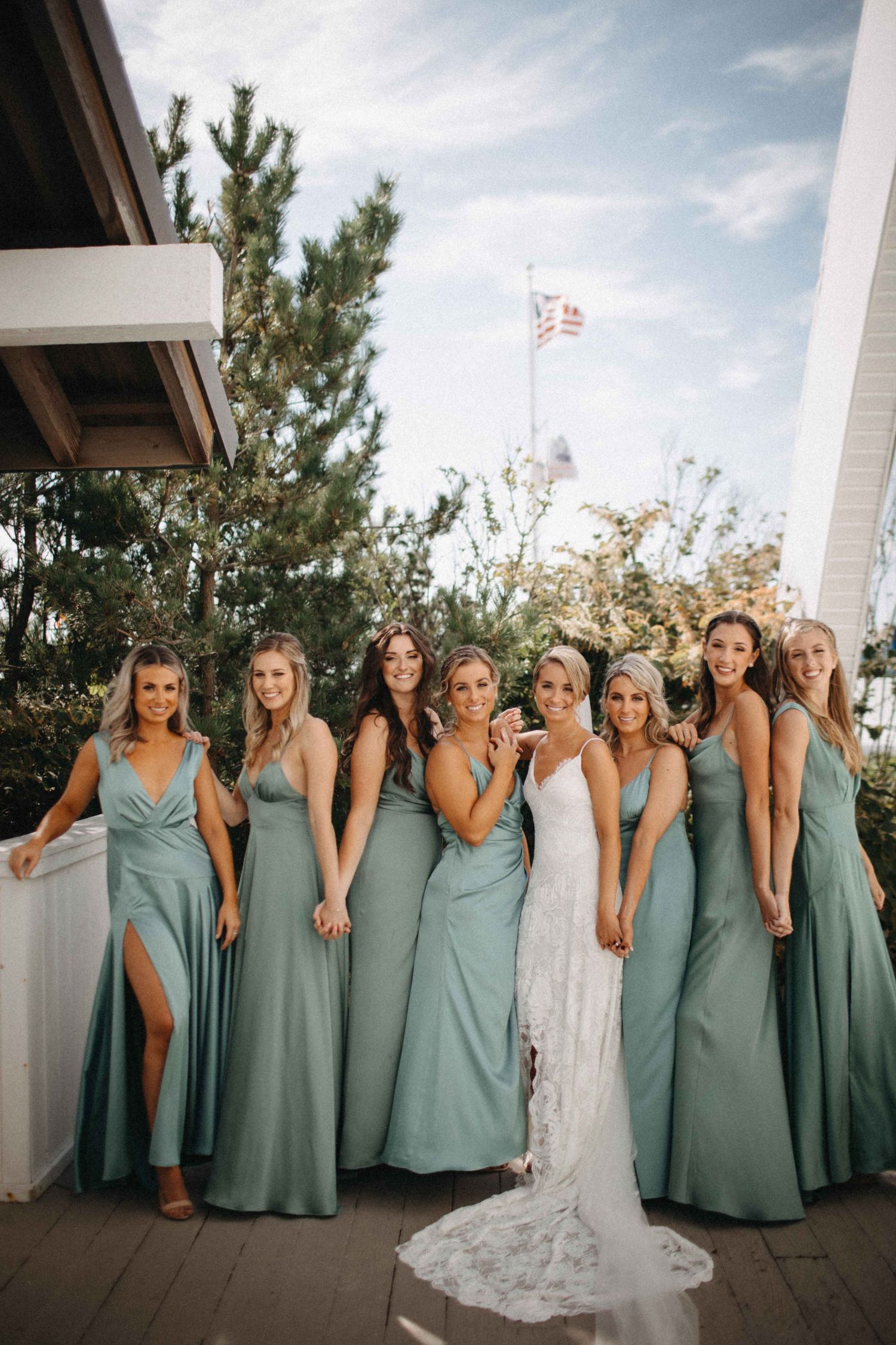 Bride poses with seven bridesmaids wearing sage green gowns on outdoor venue balcony.