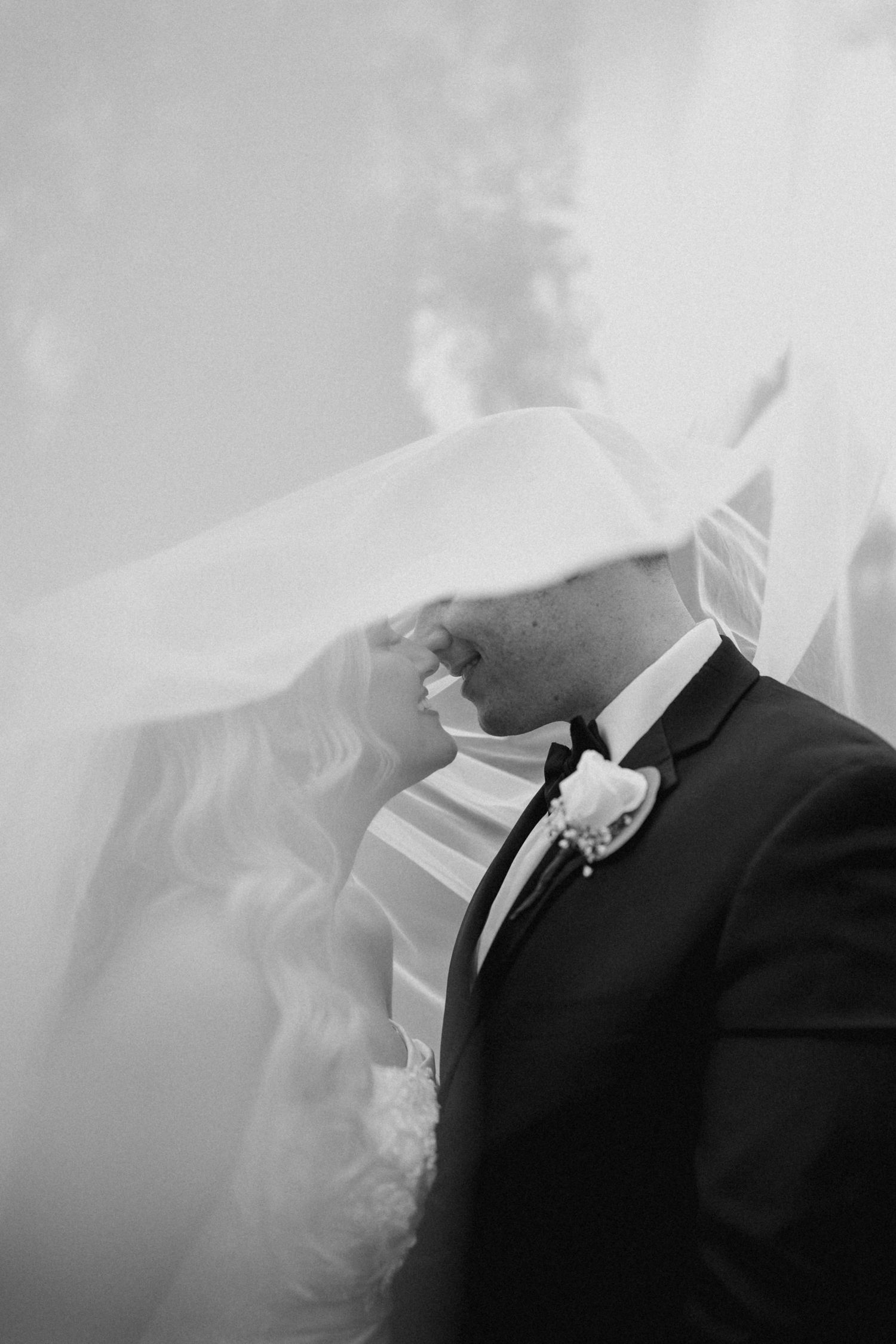 Black and white wedding photo of bride and groom sharing a kiss under flowing veil.