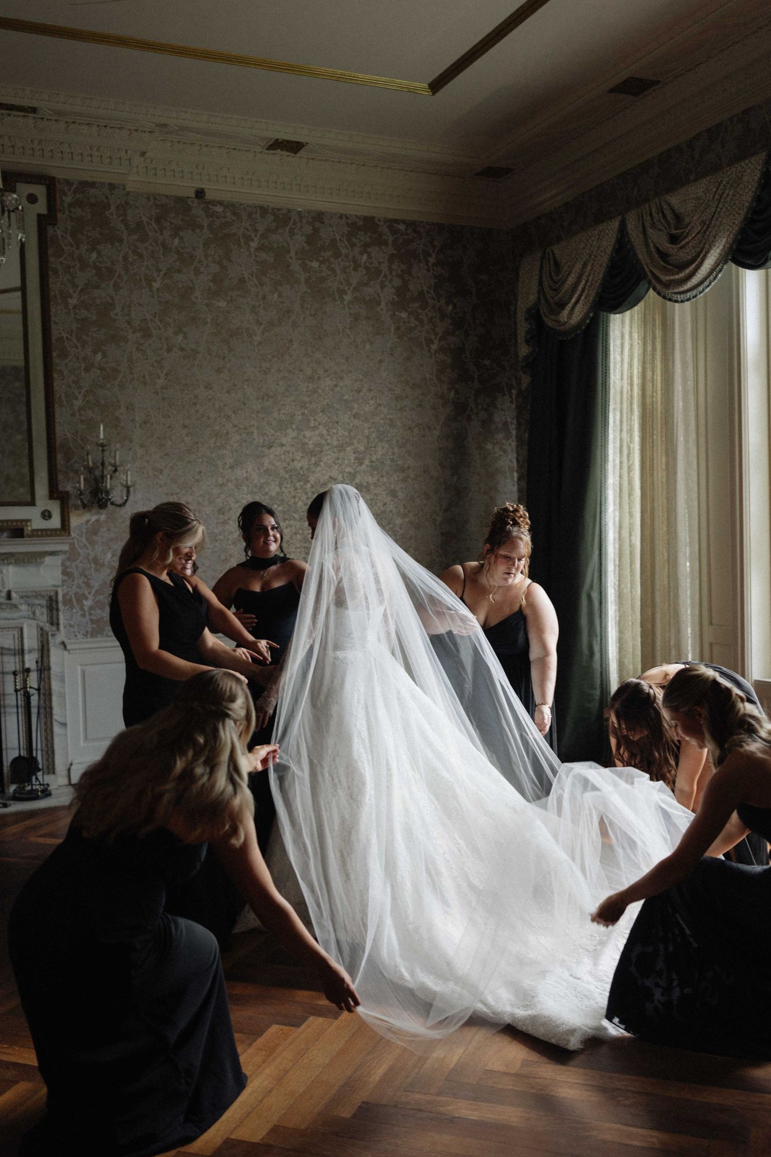 Bridesmaids in black dresses help a bride with her flowing white veil in an elegant room before the wedding.
