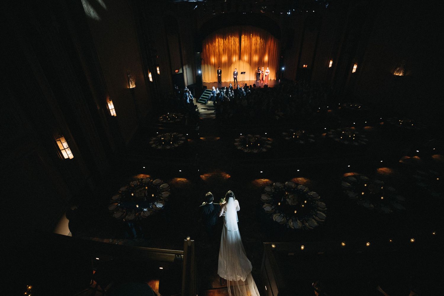 Aerial view of bride walking down aisle in grand theater wedding venue with round tables.
