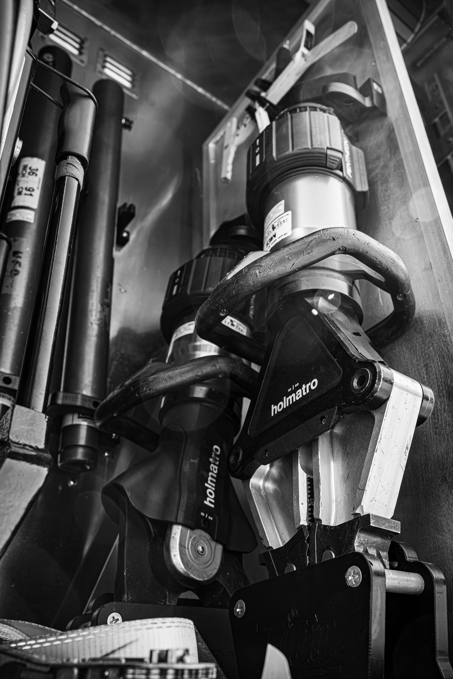 Black and white close-up of Holmatro hydraulic rescue tool stored in a fire truck compartment.
