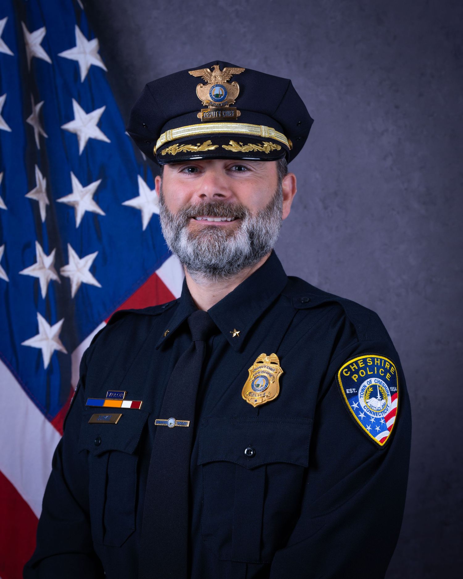 Bearded police officer in formal dark uniform with gold badge and decorated cap poses by American flag.
