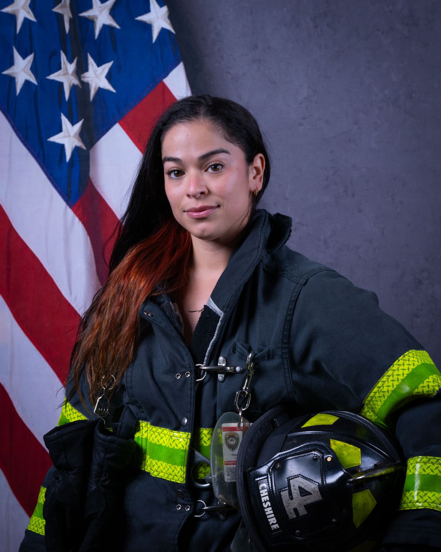 Female Cheshire firefighter in turnout gear holds helmet before American flag.