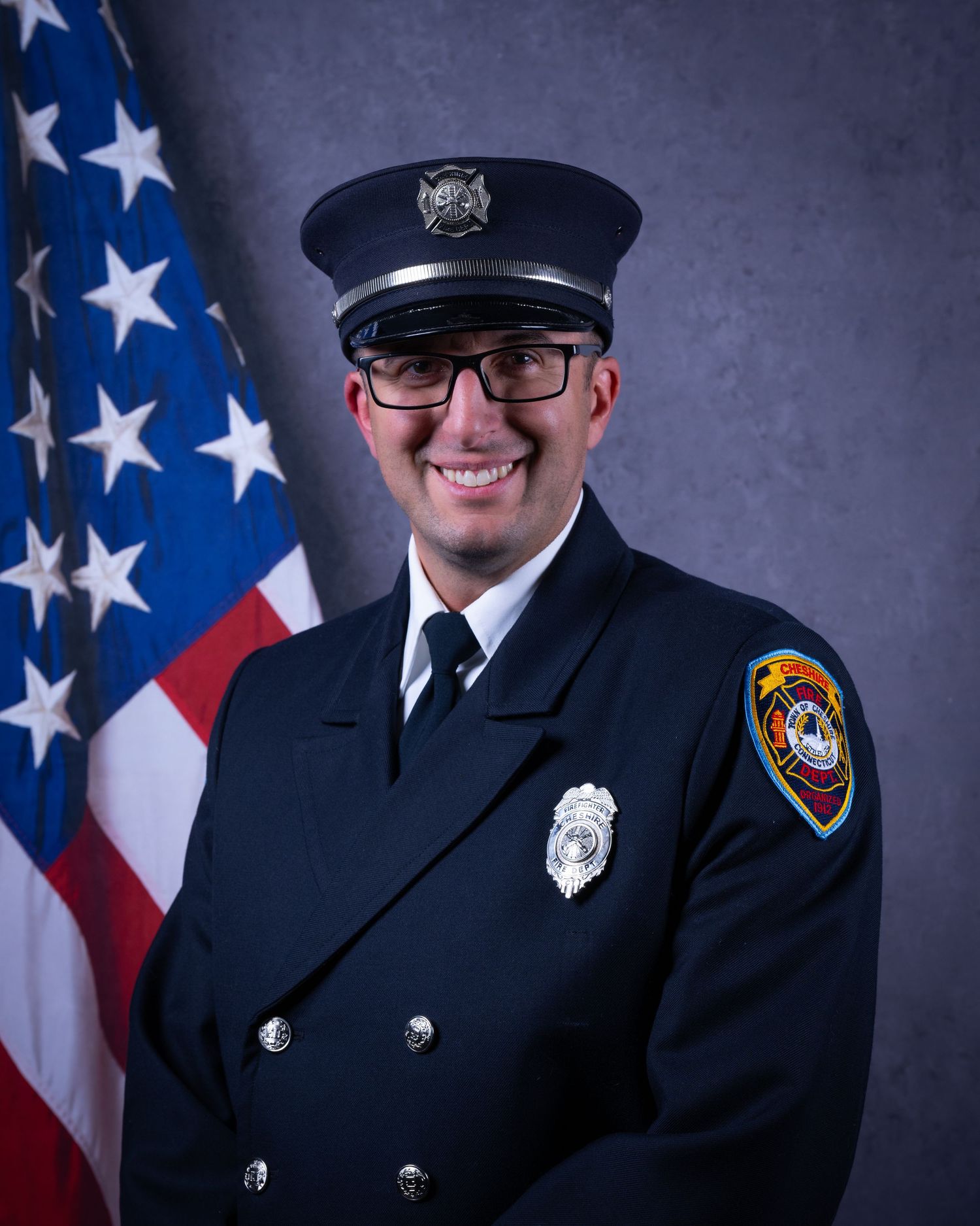 Smiling firefighter wearing glasses in formal dark dress uniform with badge poses before American flag.