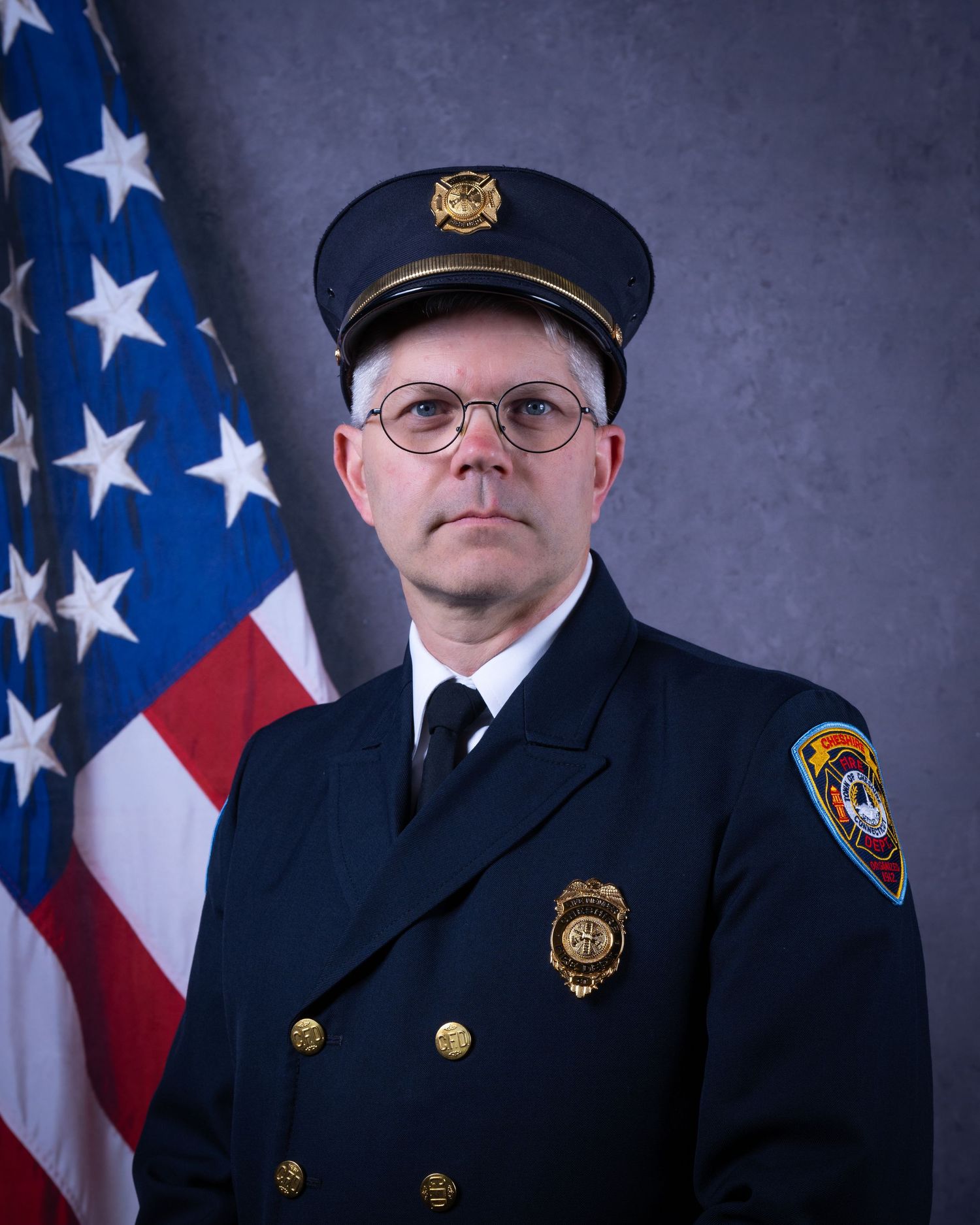 Senior firefighter in dress uniform with gold badge poses before American flag.