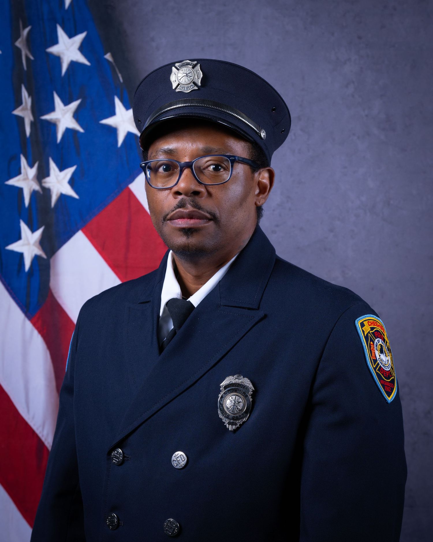 African American firefighter in formal dark dress uniform with badge and glasses poses by American flag.
