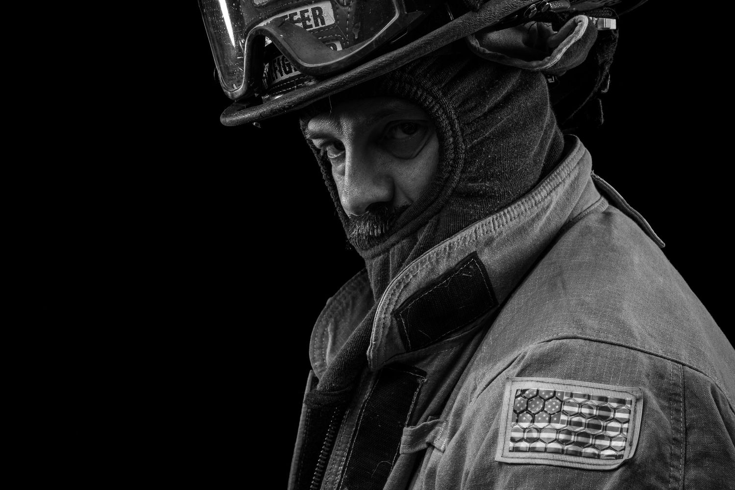 Black and white dramatic portrait of a firefighter in helmet and turnout gear on dark background.