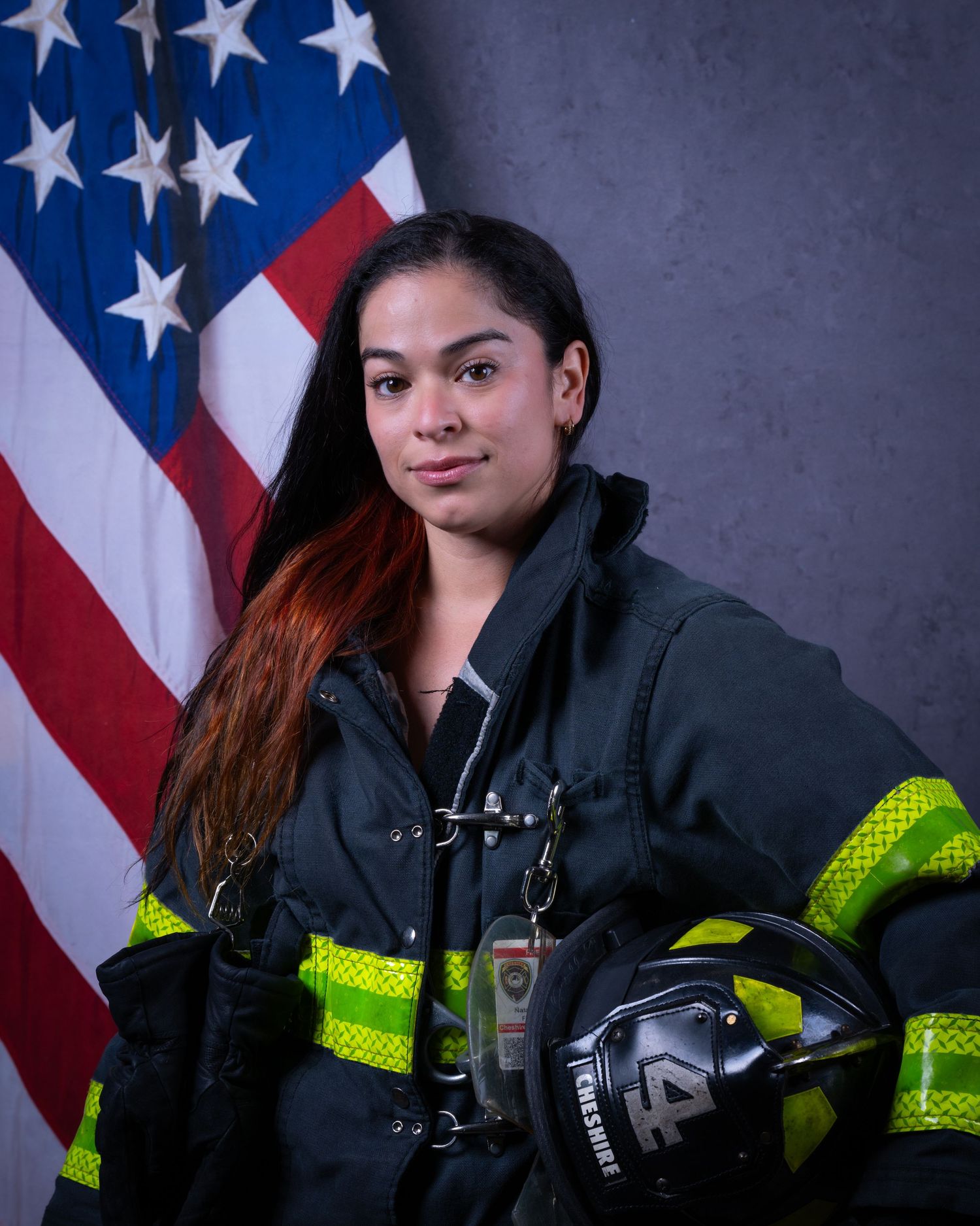 Female firefighter in turnout gear holds helmet before American flag.