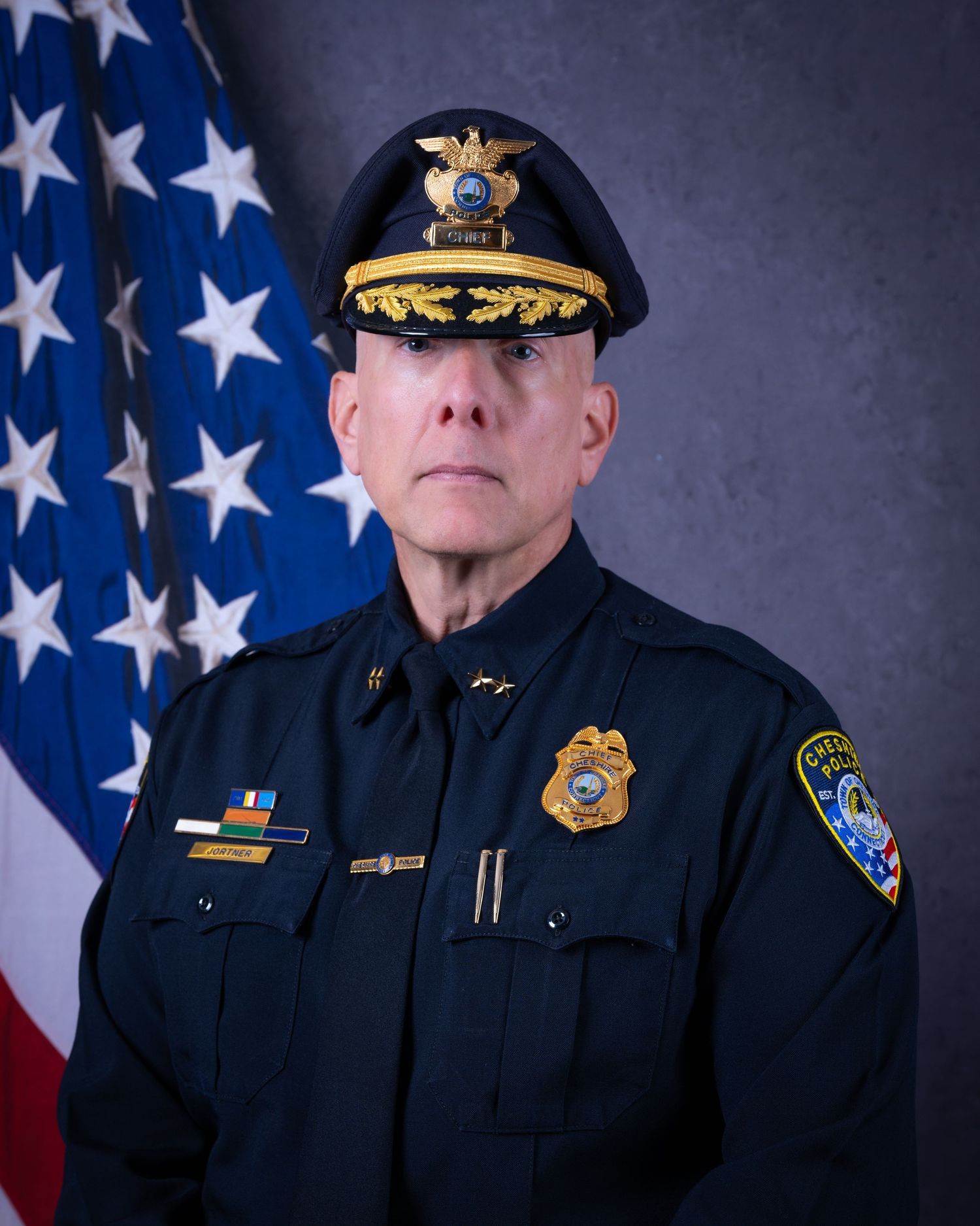 Senior police chief in formal black uniform with gold badge and decorated cap stands before American flag.