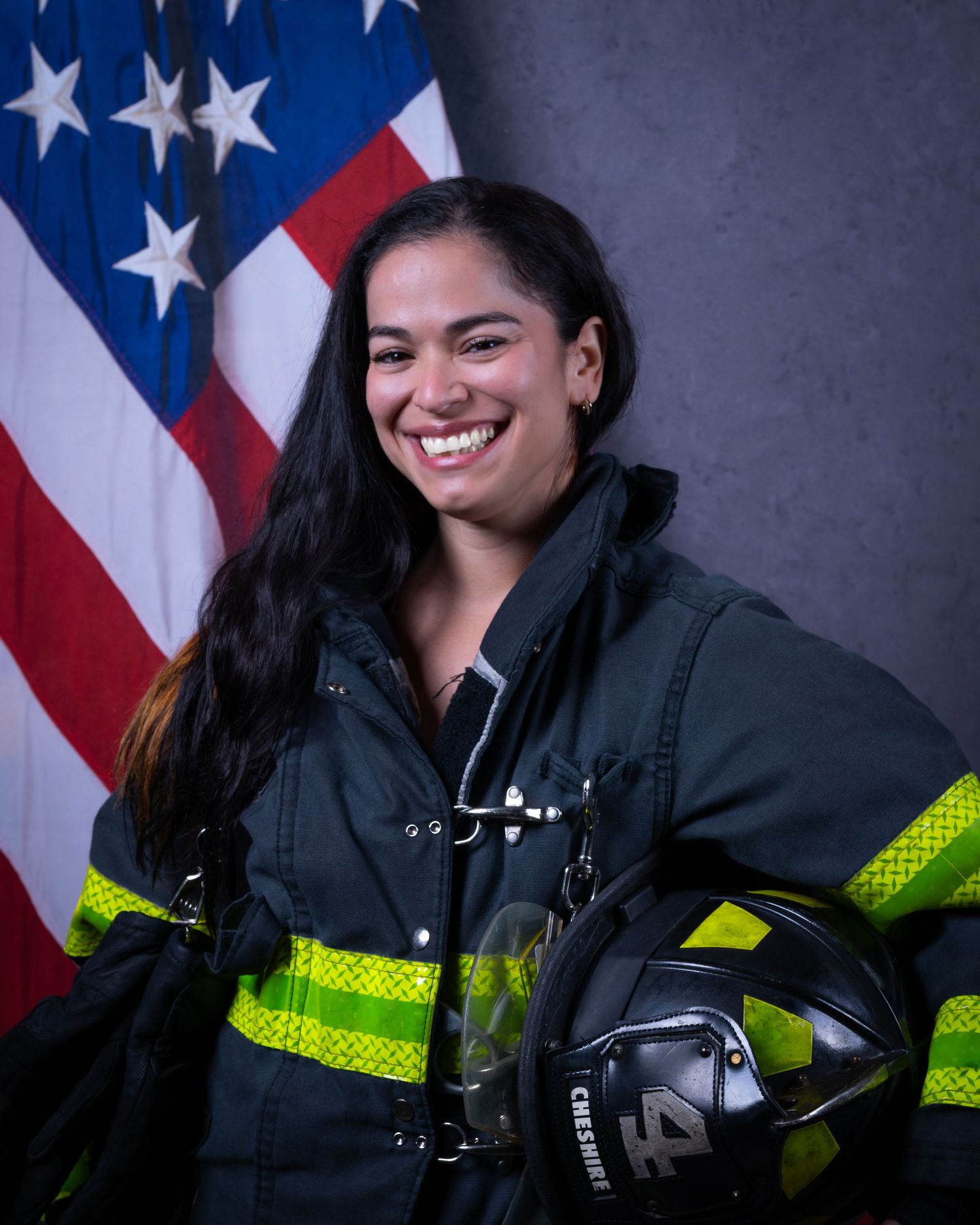 Smiling female Cheshire firefighter in turnout gear holds helmet before flag.