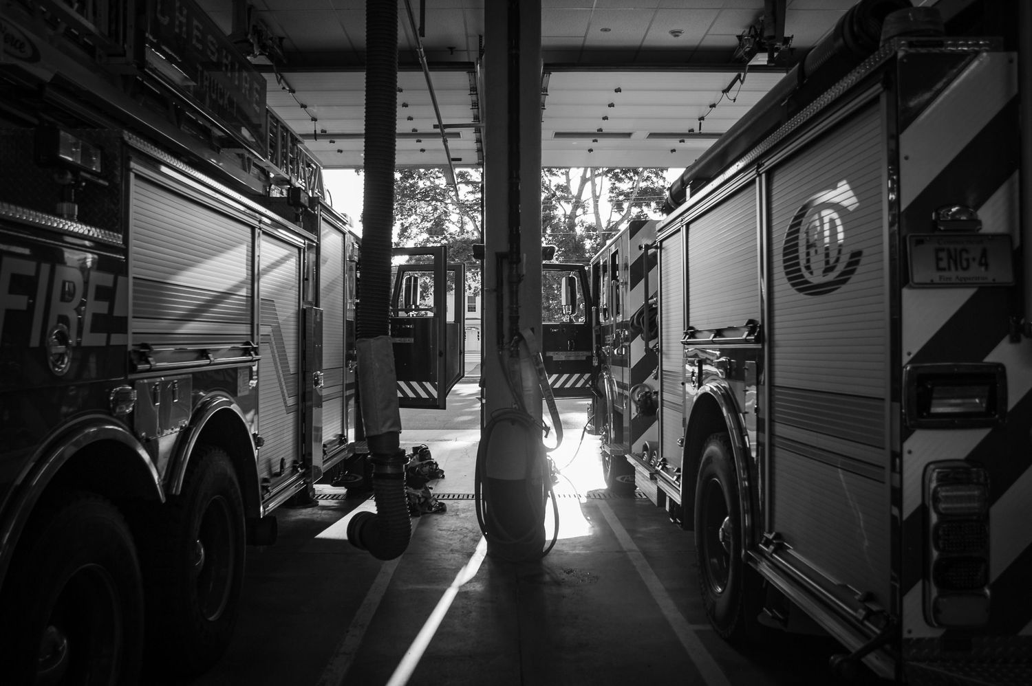 Black and white photo of two fire trucks parked inside a fire station garage with light streaming between them.