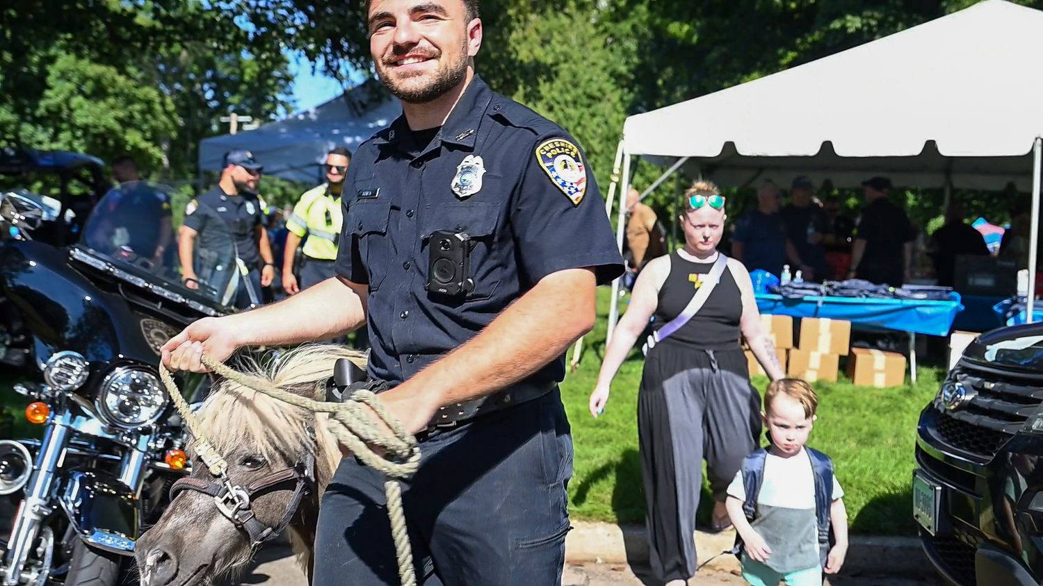 A smiling police officer holds rope near a motorcycle at an outdoor community event with families in the background.