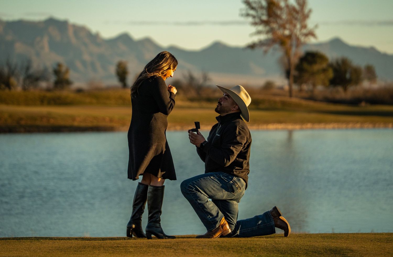 A couple sits together on a bench by a serene lake at sunset with mountains in the background.