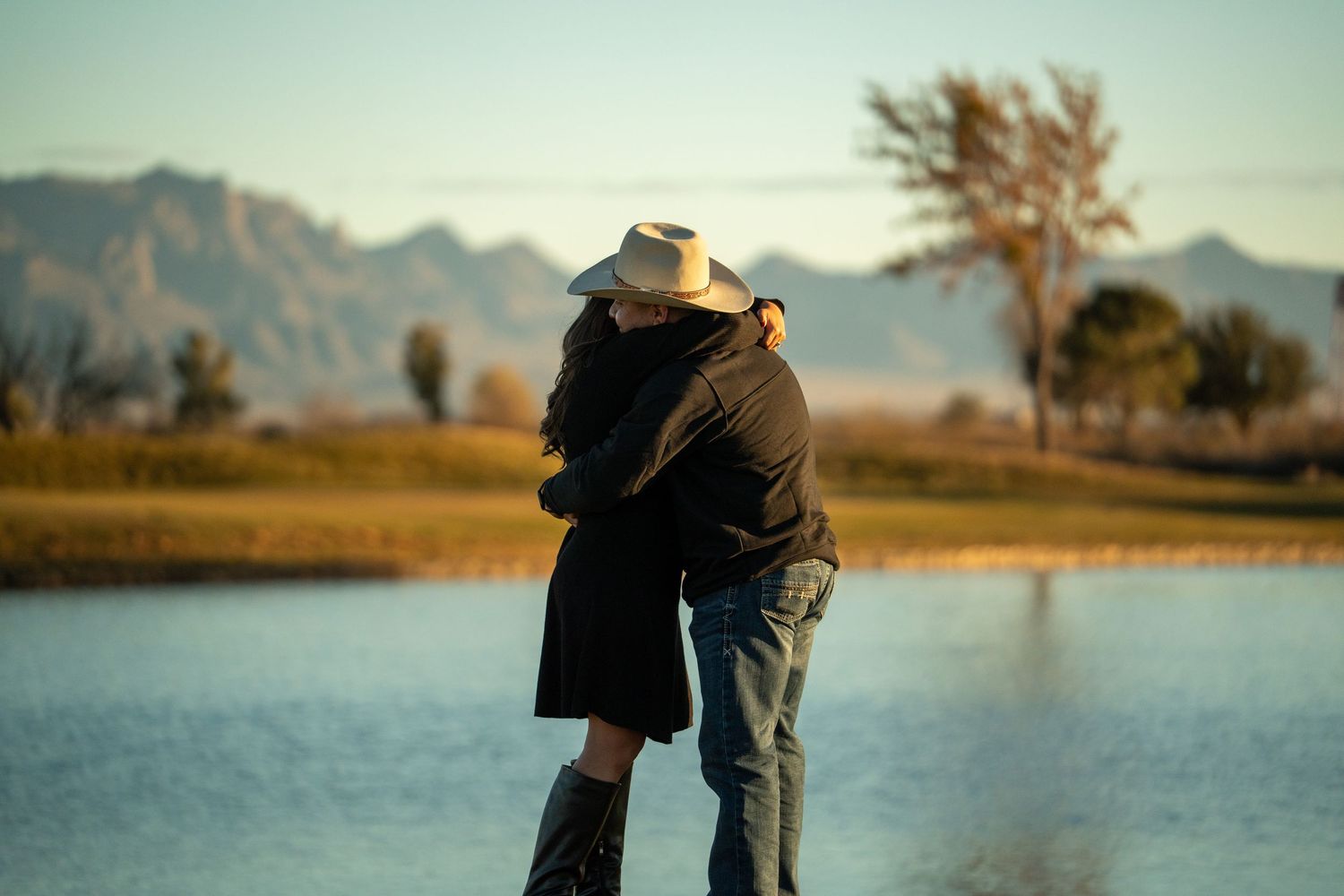 A couple embraces while standing by a serene lake with mountains and a lone tree in the background at sunset.