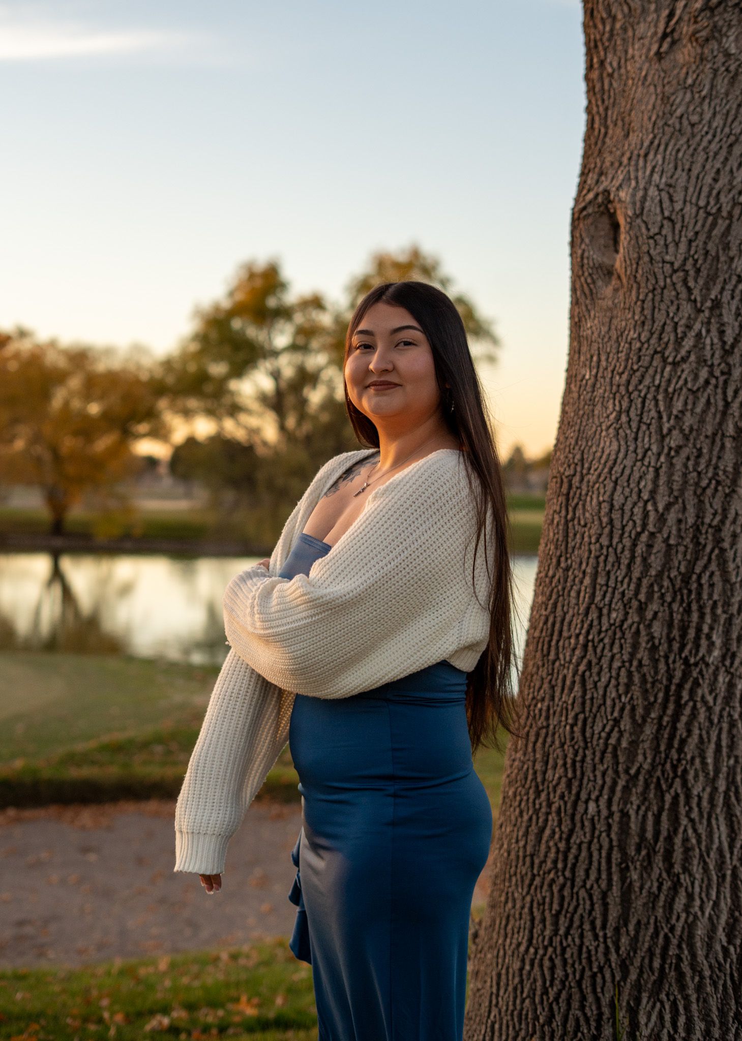Person in cream sweater and blue dress poses against tree trunk by lake at sunset.